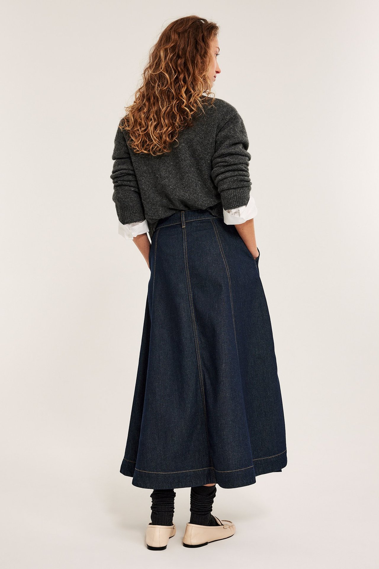 Bakfra: kvinne i mørkt A-linje-jeanskjørt med lommer, mørkegrå genser, hvit skjorte og beige loafers.