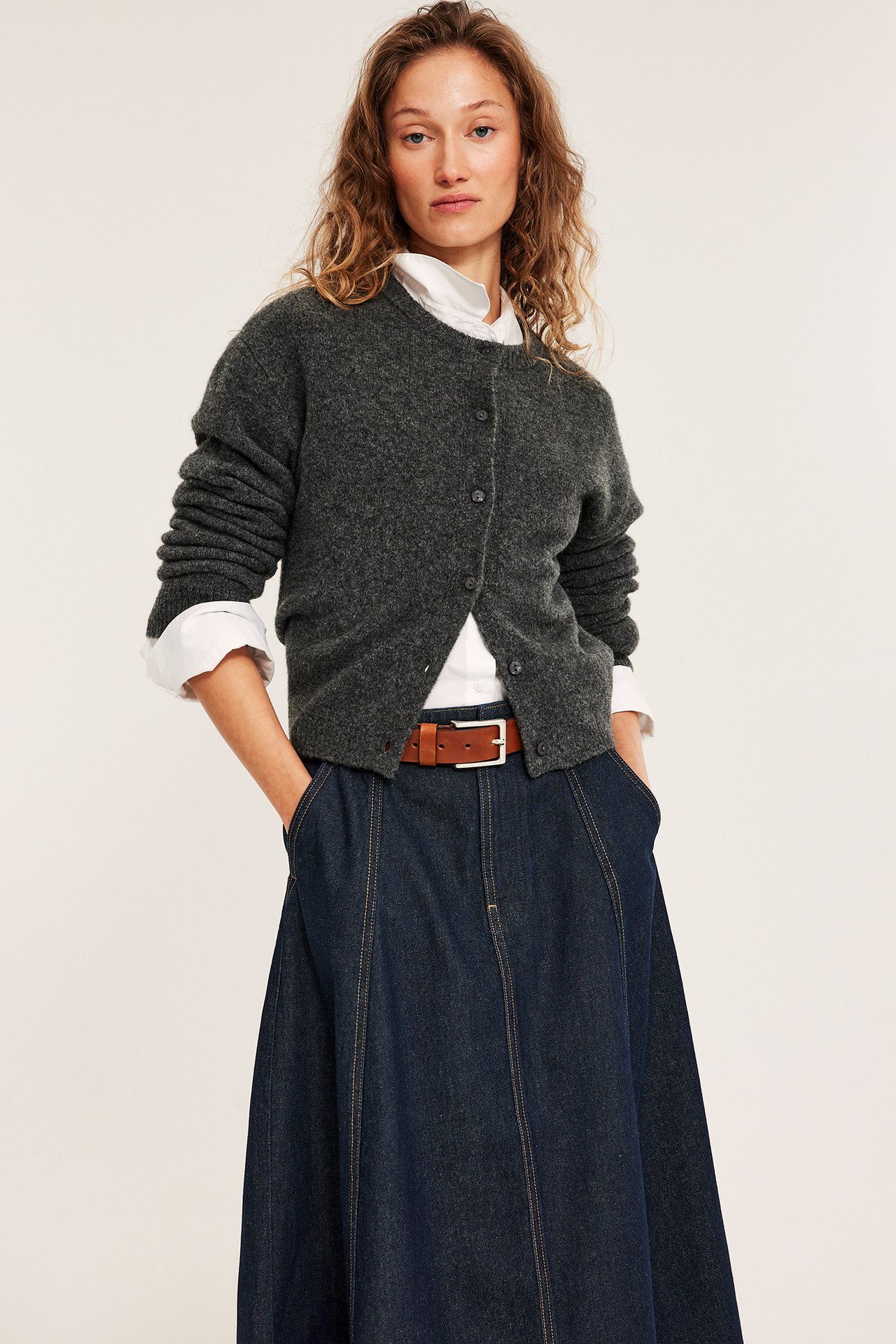 Frontvisning av kvinne i mørkblå A-linje denimskjørt, grå cardigan, hvit skjorte og brunt belte.