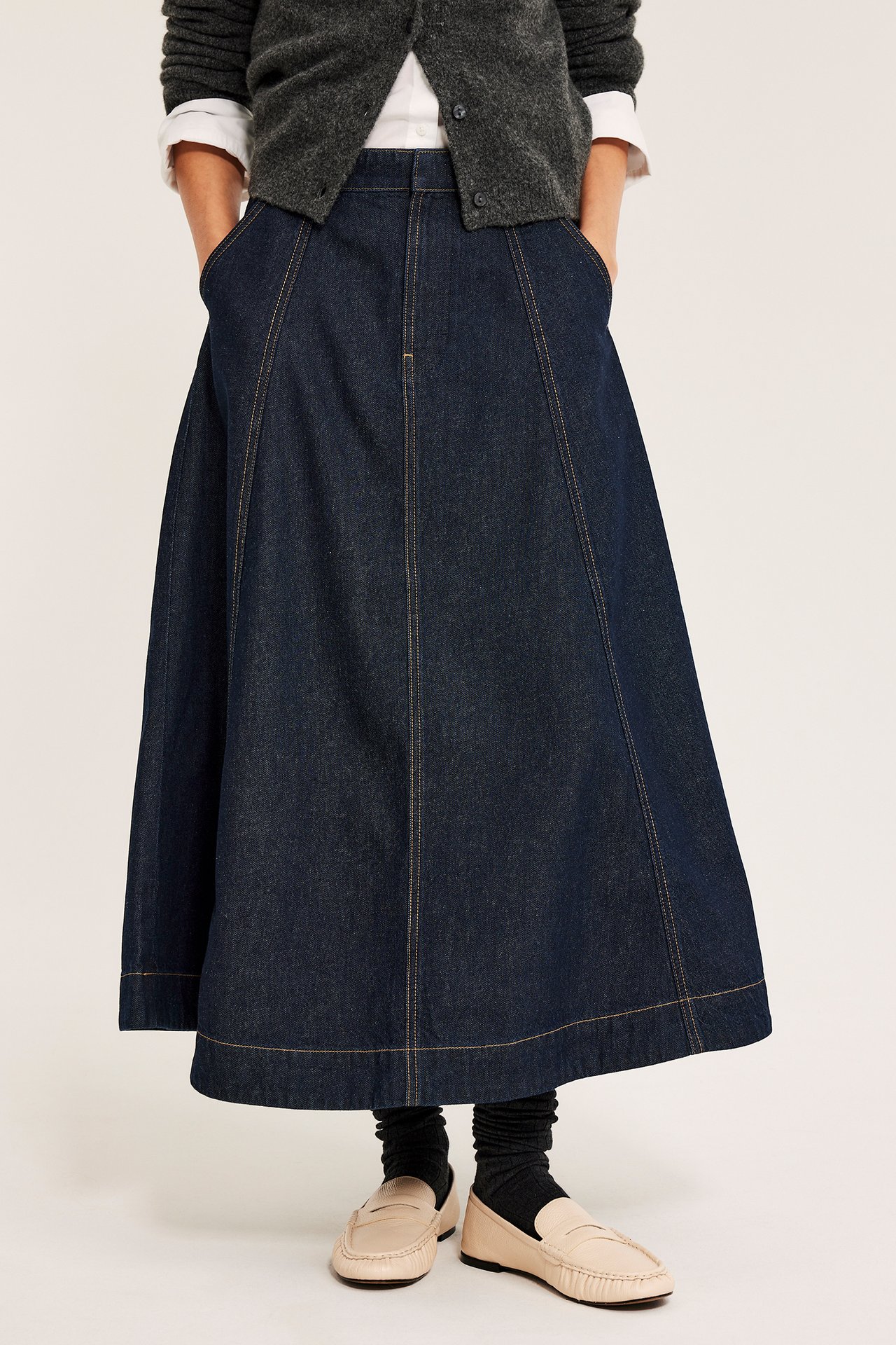 Frontbilde: kvinne i mørkt A-formet dongeriskjørt med sidelommer, grå cardigan, hvit skjorte og beige loafers.