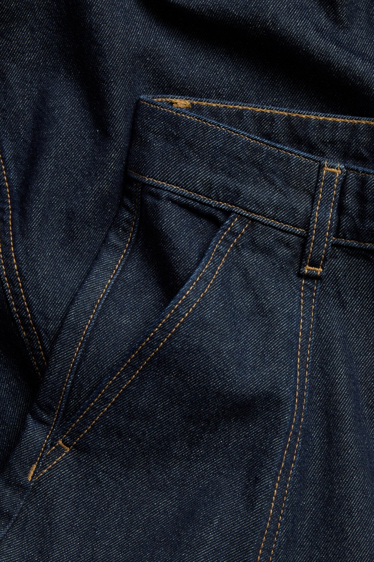 Nærbilde av et mørkeblått langt A-formet dongeriskjørt med sidelomme og kontrastsømmer.