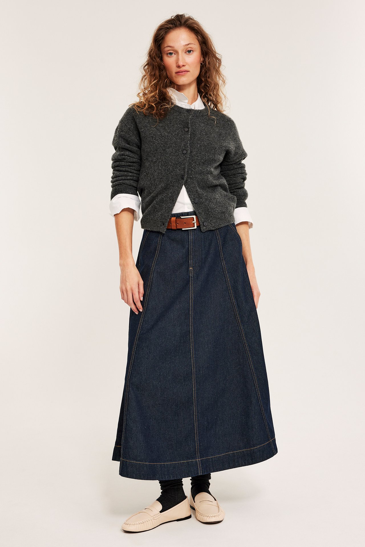 Forfra: Kvinne i mørkt denim A-linjeskjørt, mørkegrå cardigan, hvit skjorte, beige loafers.