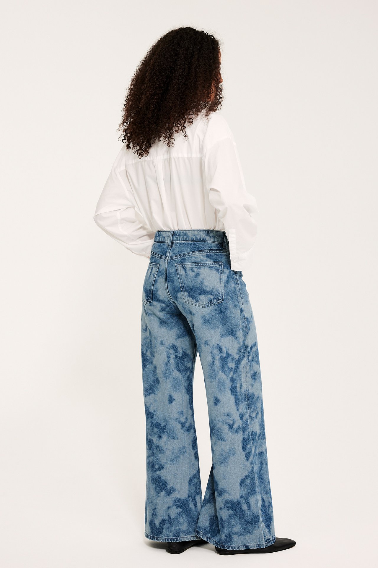 Takaapäin: nainen vaaleansinisissä leveissä korkeavyötäröisissä batikfarkuissa, valkoisessa paidassa ja mustissa kengissä.