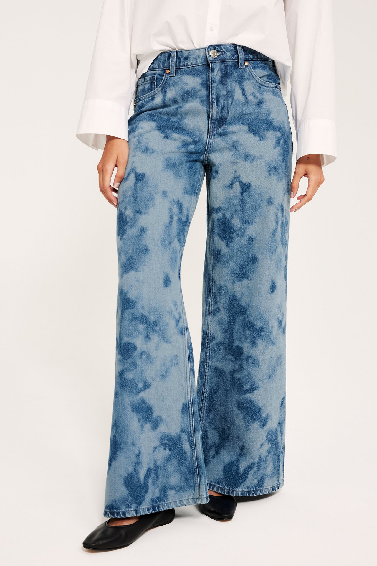 Naisen etukuva vaaleissa, valkaistuissa, korkeavyötäröisissä leveälahkeisissa farkuissa, valkoisessa paidassa ja mustissa kengissä.