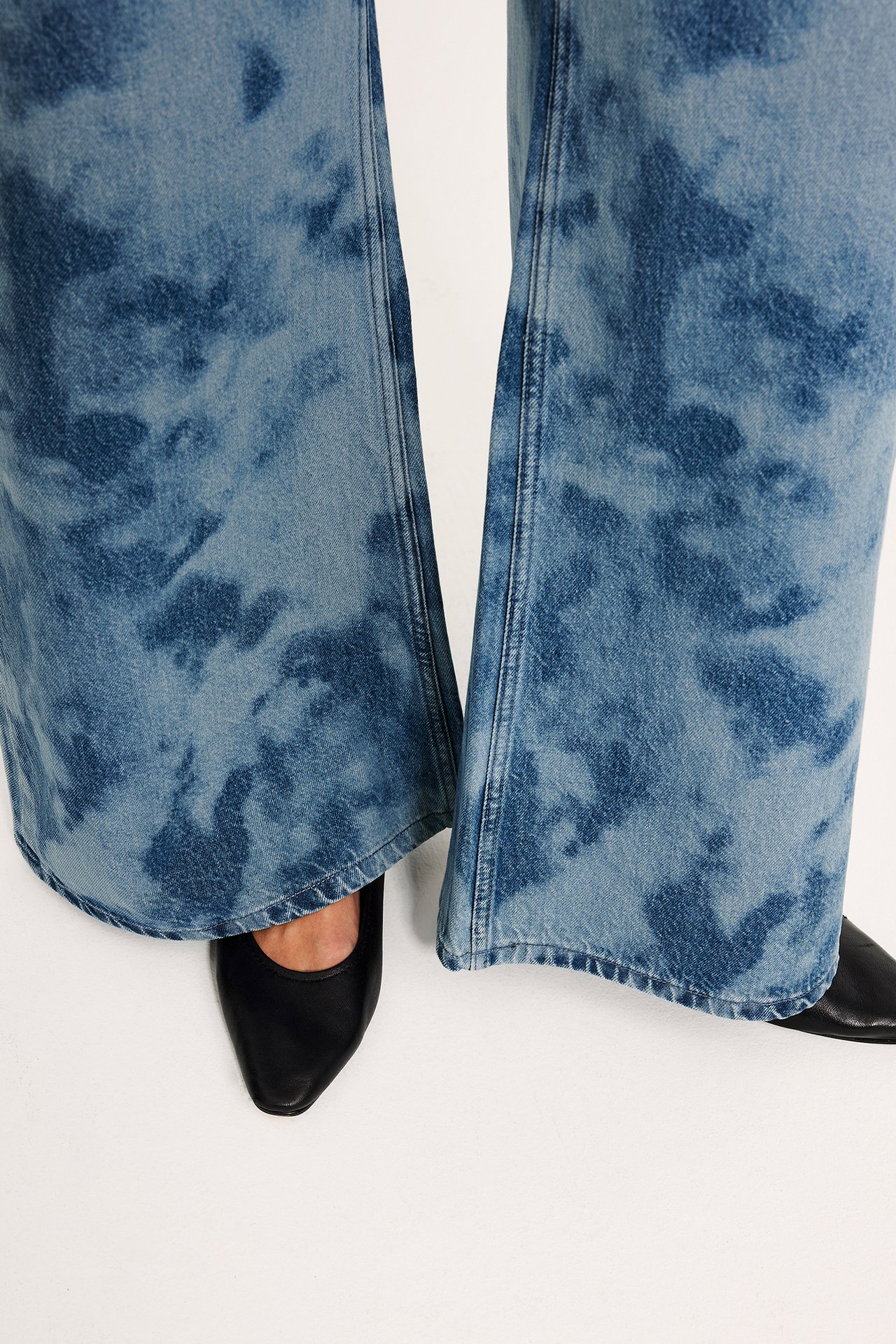 Lähikuva naisen jaloista, yllään vaaleansiniset batikkikuvioidut leveät farkut ja mustat kengät.