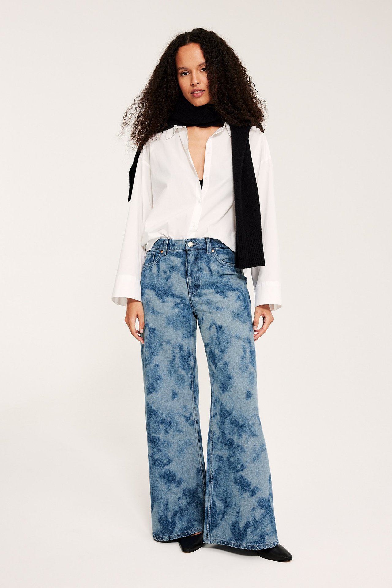 Edestä, nainen vaaleansinisissä batikkuvioisissa leveissä farkuissa, valkoisessa paidassa ja mustassa huivissa.