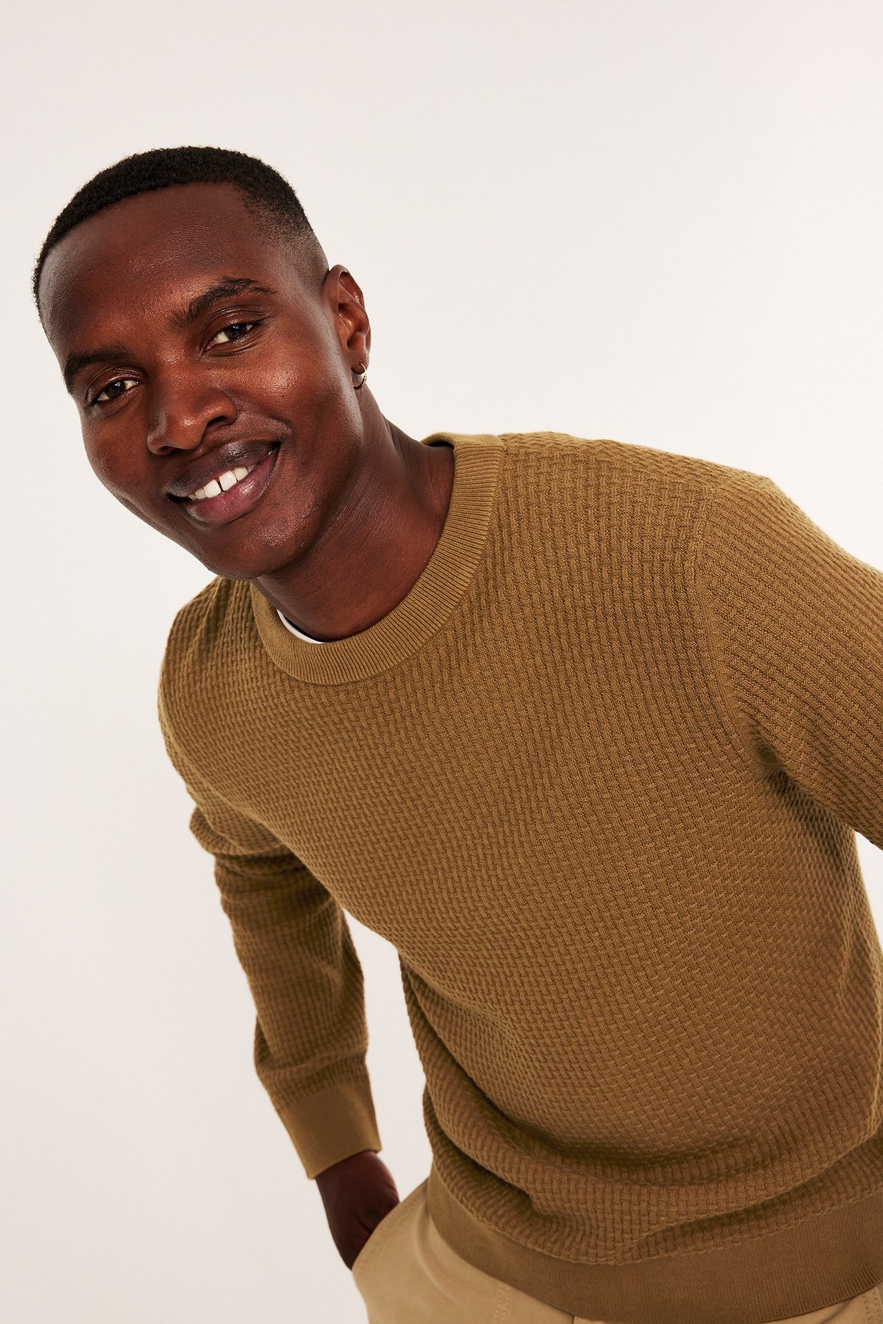 Framifrån: Leende man i en brun strukturstickad bomullströja och ljusbeige byxor.