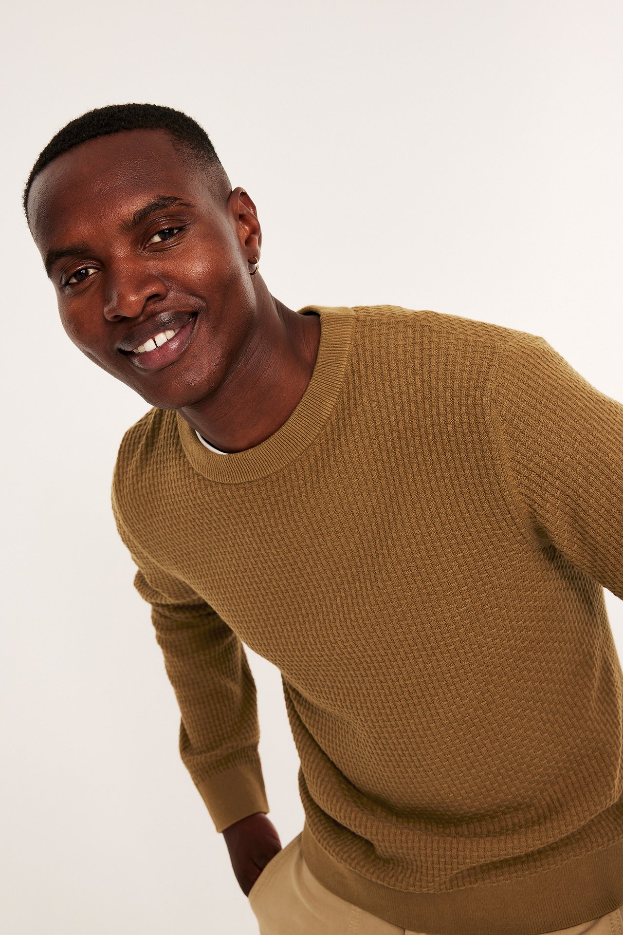 Framifrån: Leende man i en brun strukturstickad bomullströja och ljusbeige byxor.