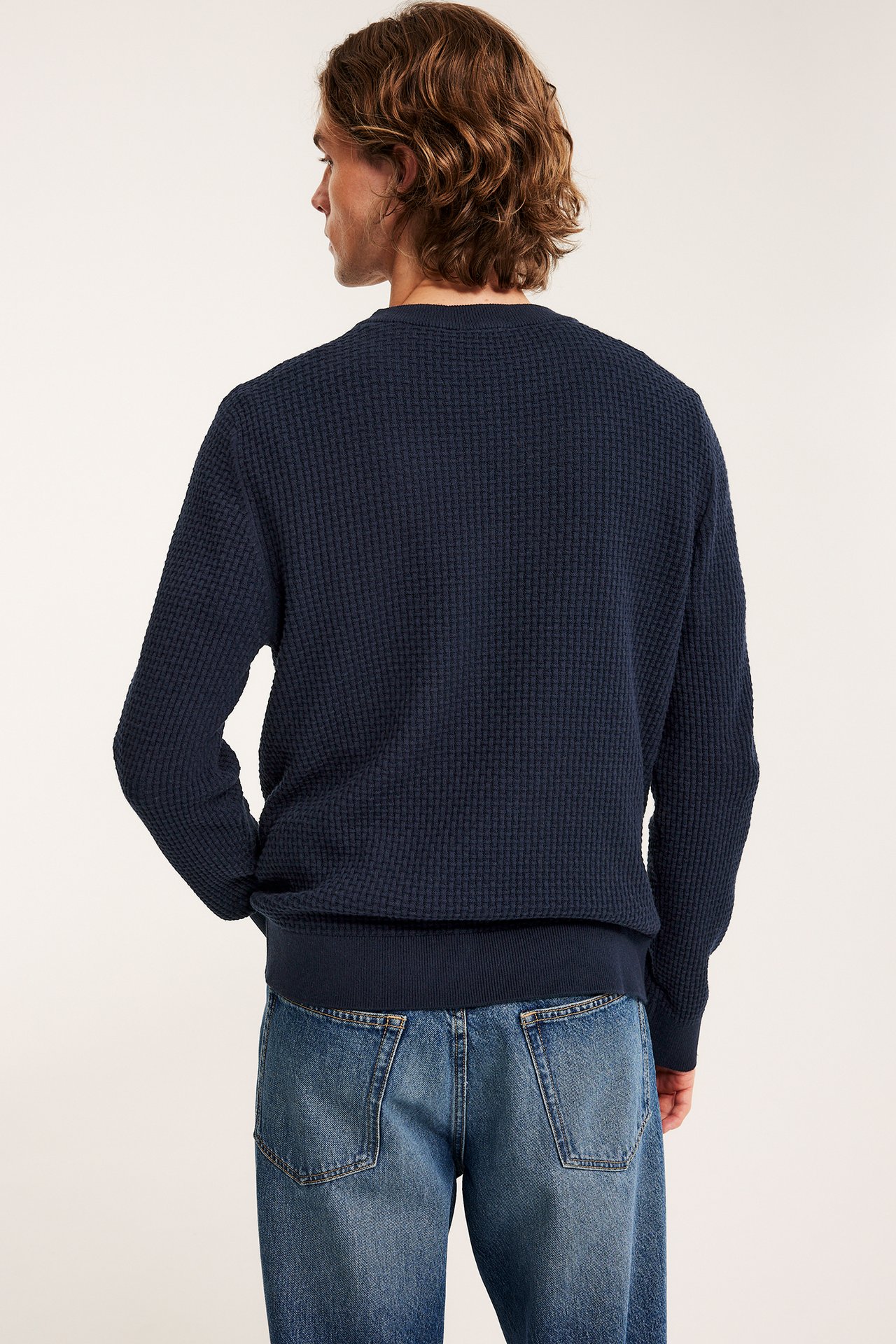 Bakfra på en mann i en mørk blå strukturstrikket bomullsgenser og blå vide jeans.