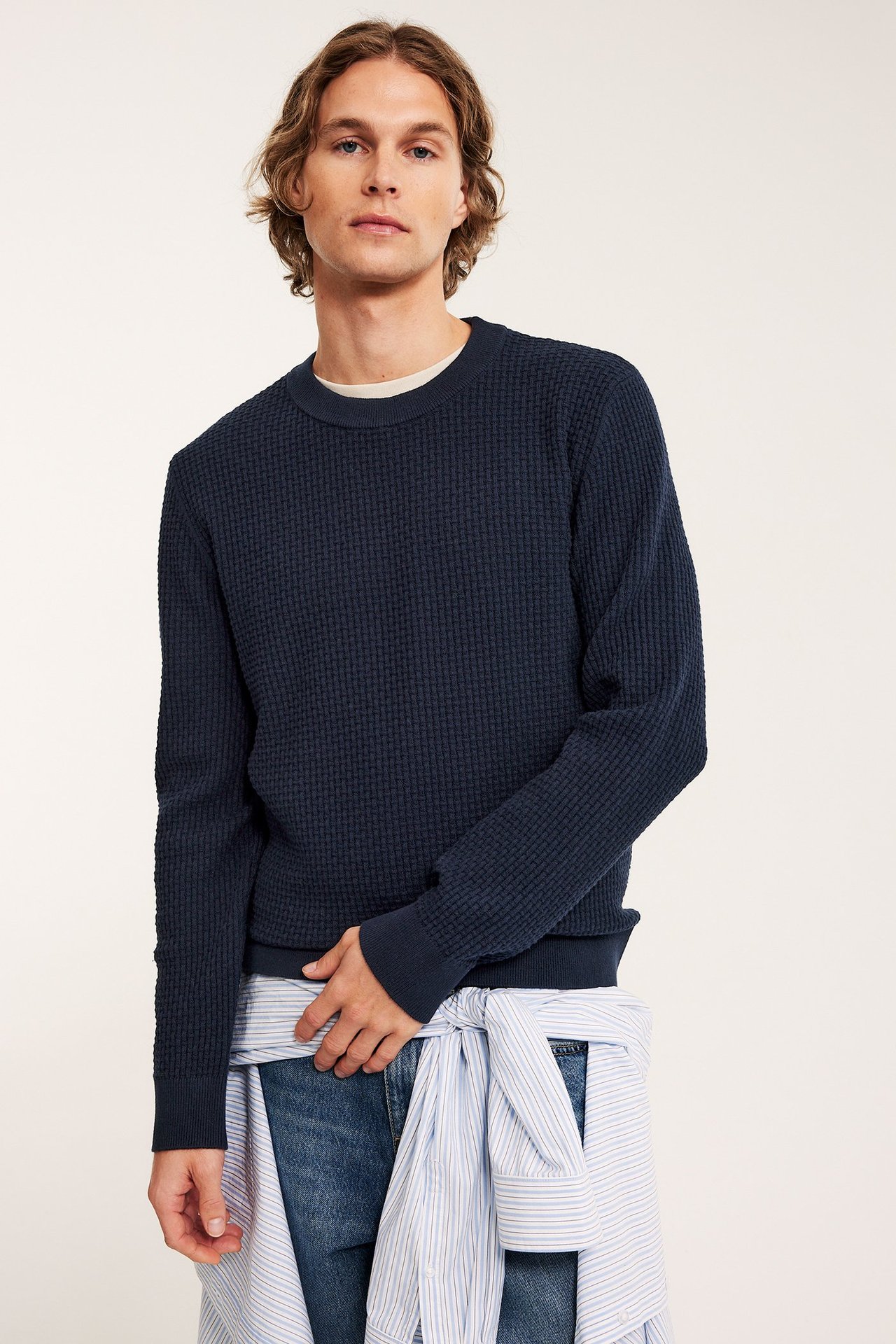Frontbilde: Mann i mørkeblå strukturstrikket bomullsgenser, lys t-skjorte, blå jeans og stripet skjorte knyttet i midjen.