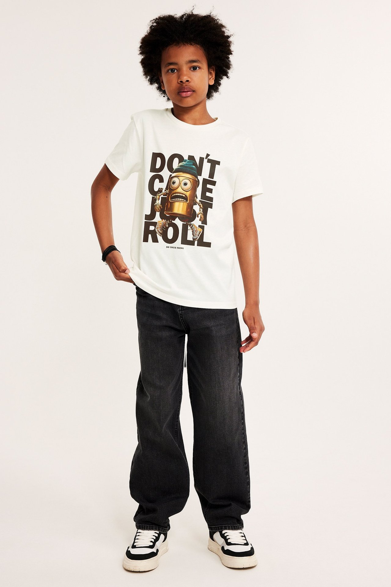 Widok z przodu: dziecko w kremowym bawełnianym T-shircie z nadrukiem robota, ciemnoszarych jeansach i sneakersach.
