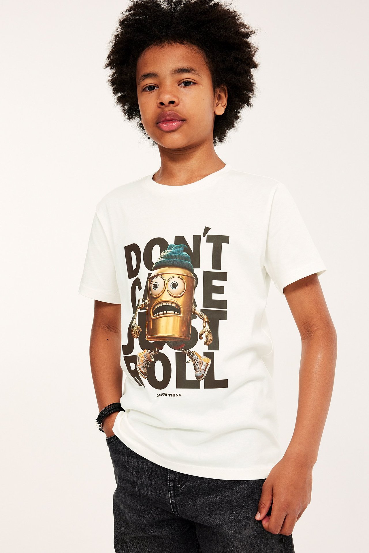 Widok z przodu: Dziecko w bawełnianym T-shircie ecru z robotem, ciemnych dżinsach i czarnej bransoletce.