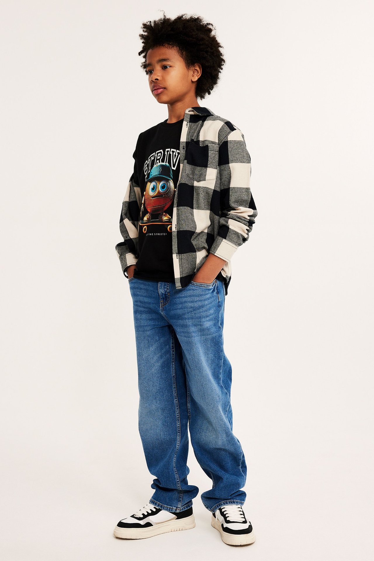 Widok z przodu: Dziecko w czarnym T-shircie z nadrukiem, otwartej koszuli w kratę, niebieskich dżinsach i sneakersach.