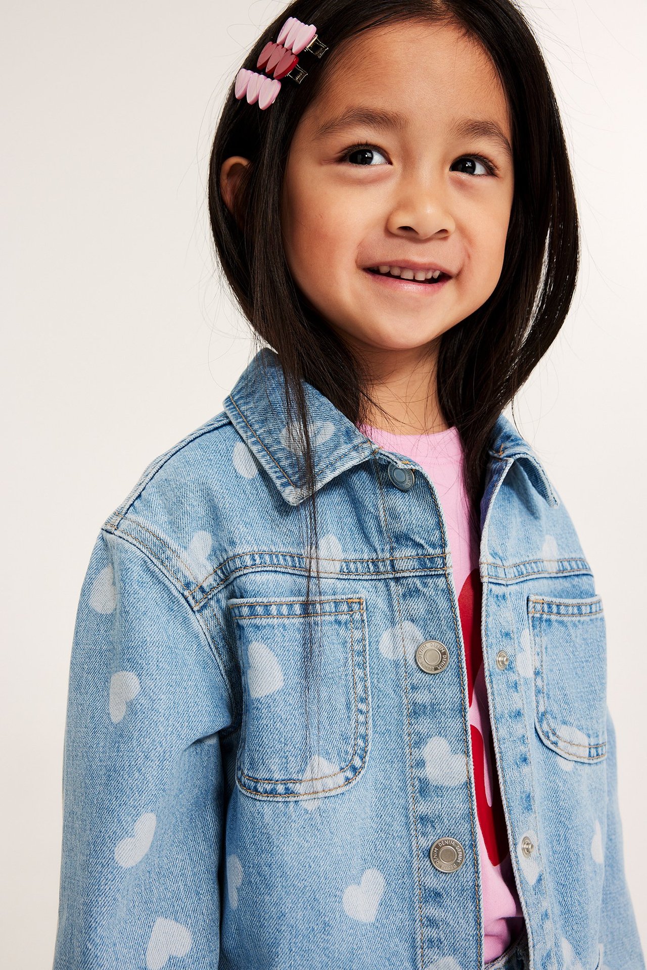 Barn bär ljusblå jeansjacka med hjärtmönster, tryckknappar, rosa t-shirt och rosa hårspännen.