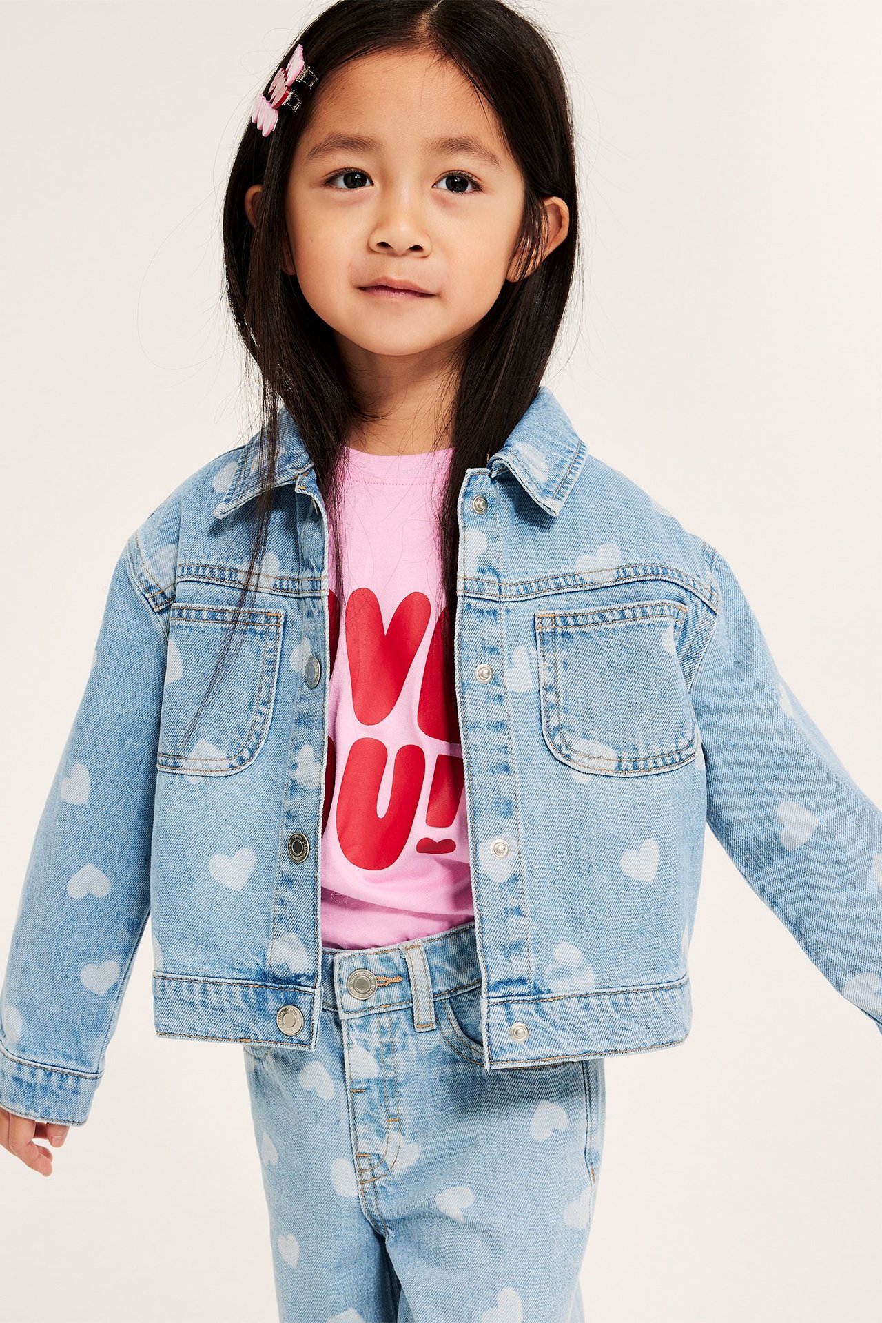 Forfra, et barn i lys dongeri hjerte-mønstret jakke med trykknapper, rosa t-skjorte og matchende jeans.
