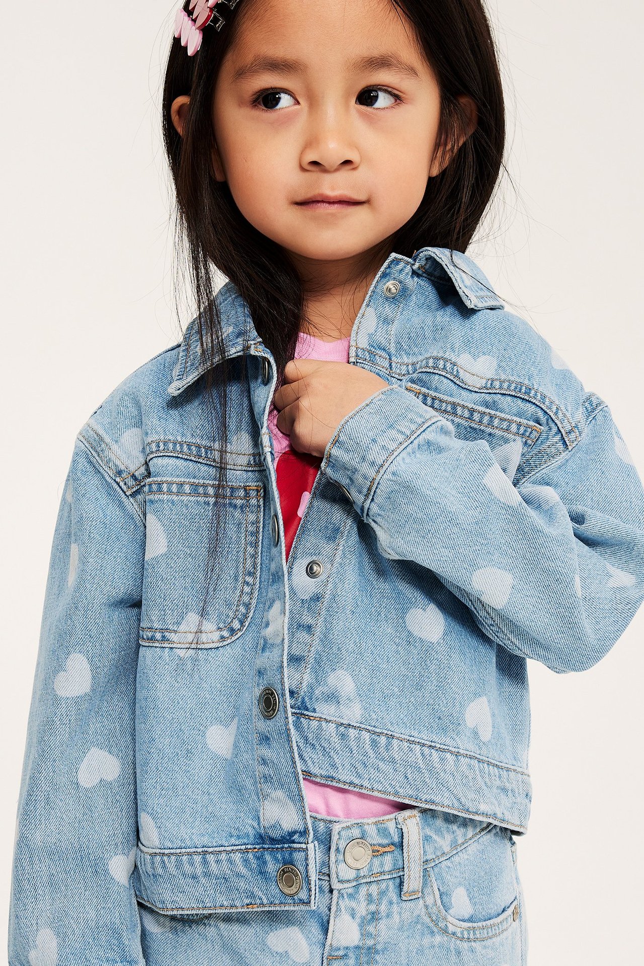 Nærbilde av barn i lys denim hjertemønstret dongerijakke med trykknapper, over rosa t-skjorte og jeans.