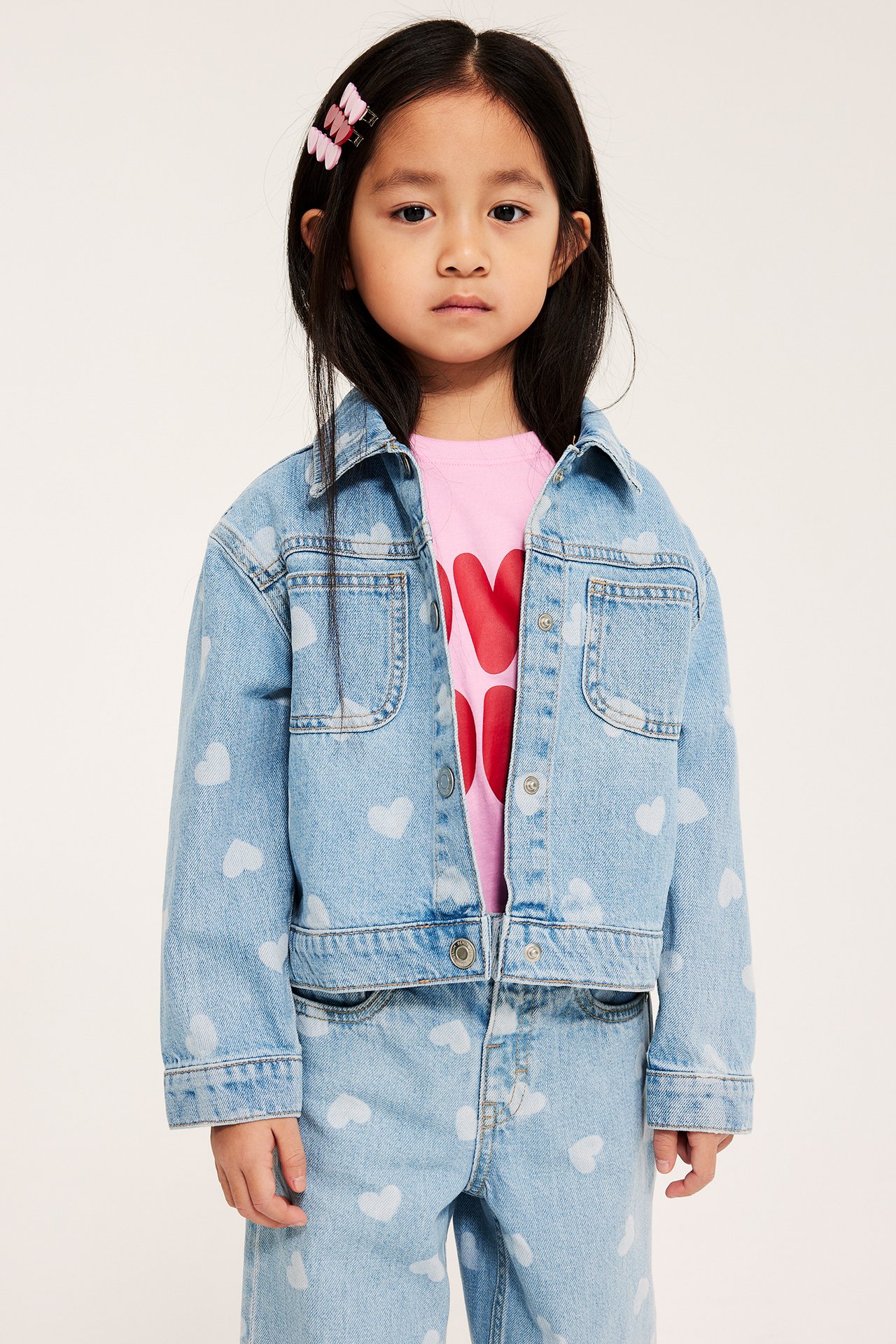 Frontalt: kid i ljusblå hjärtmönstrad jeansjacka, rosa t-shirt, matchande jeans, rosa hårklämmor.