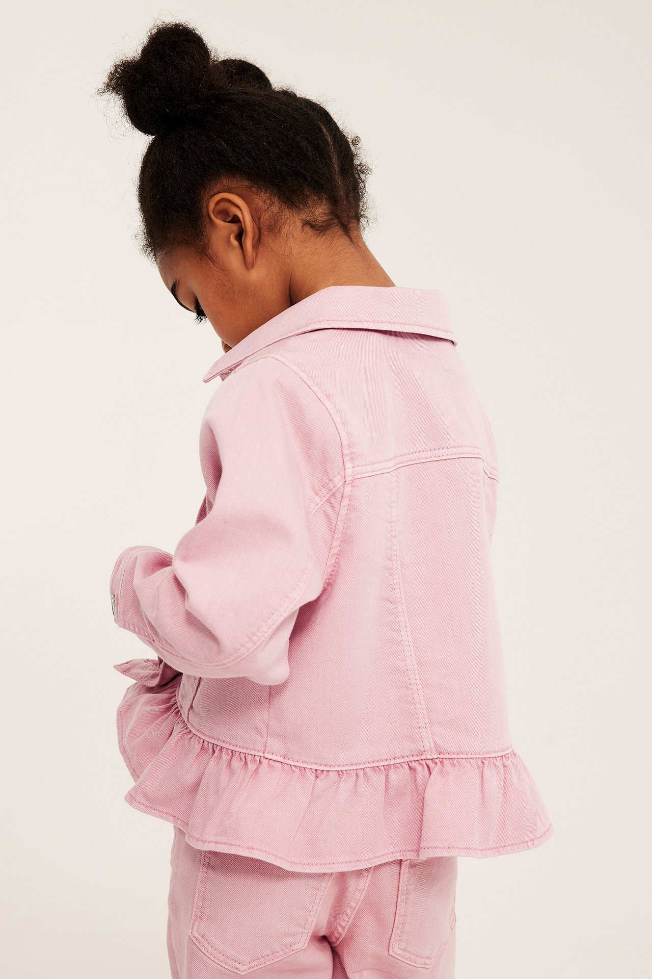 Ryggvy av ett barn i ljusrosa twill-jeansjacka med volang och matchande rosa jeans.