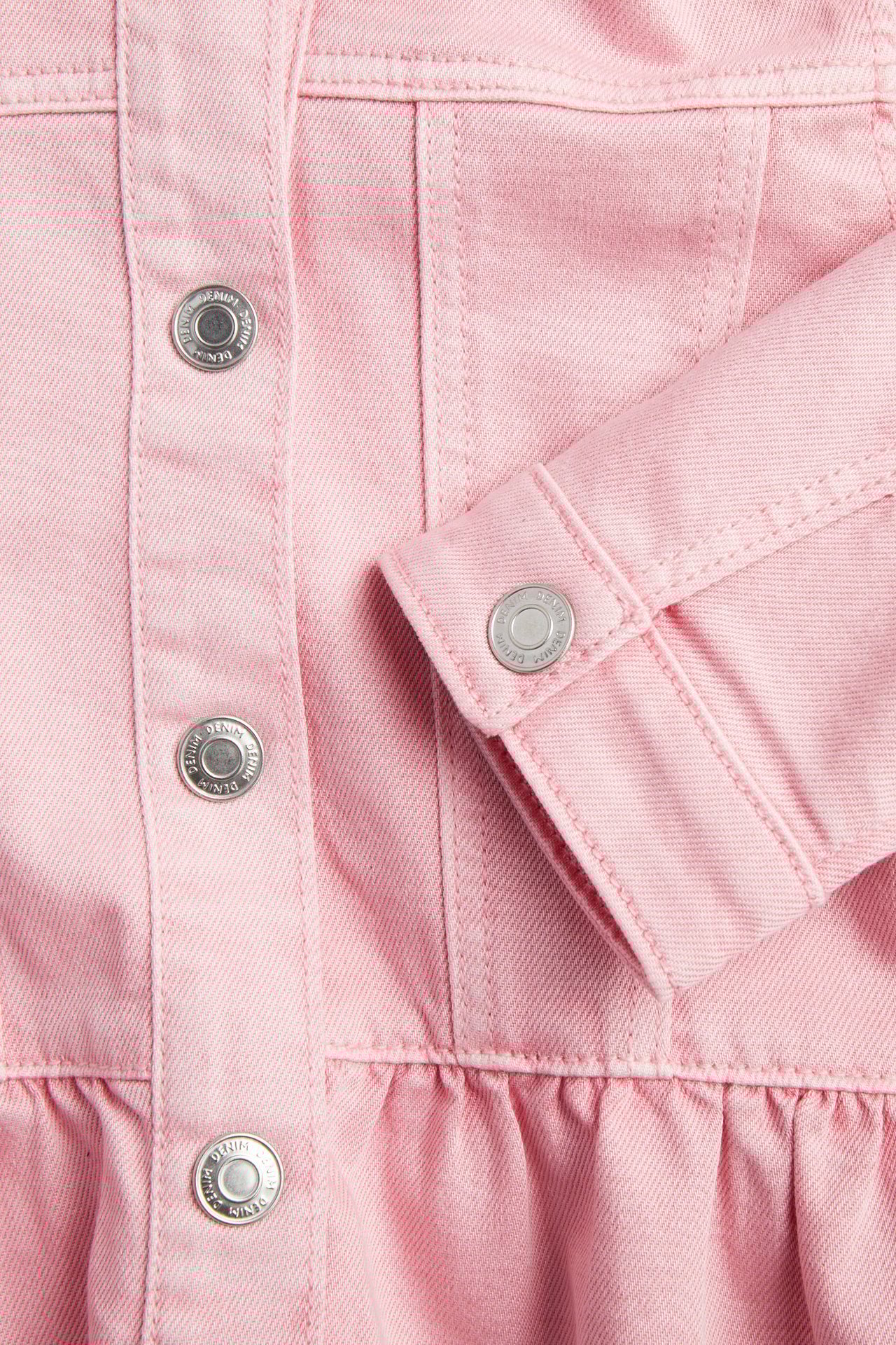Nærbilde av en lys rosa twill-dongerijakke for barn med volang og sølvfargede trykknapper.