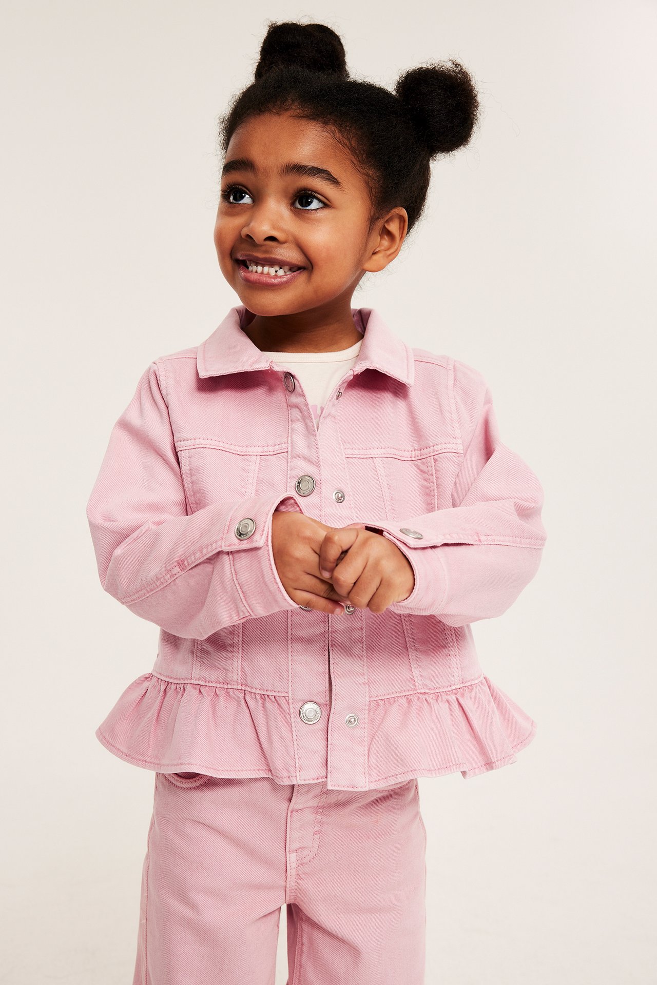 Frontvisning av et barn i en lyserosa twilljakke med volang, trykknapper og matchende bukser.