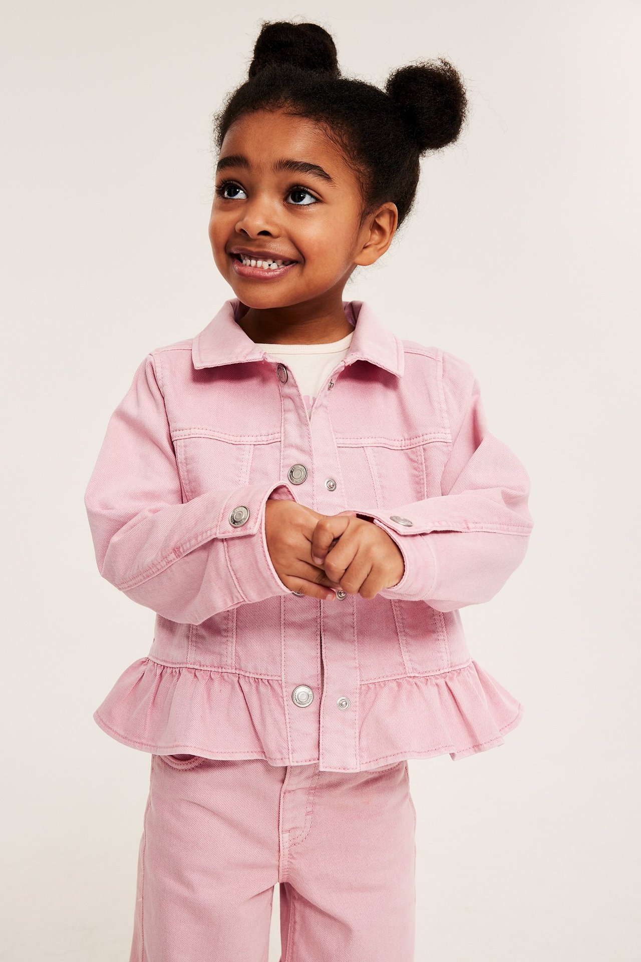 Frontvy av ett barn i en ljusrosa twilljacka med volang, tryckknappar och matchande byxor.