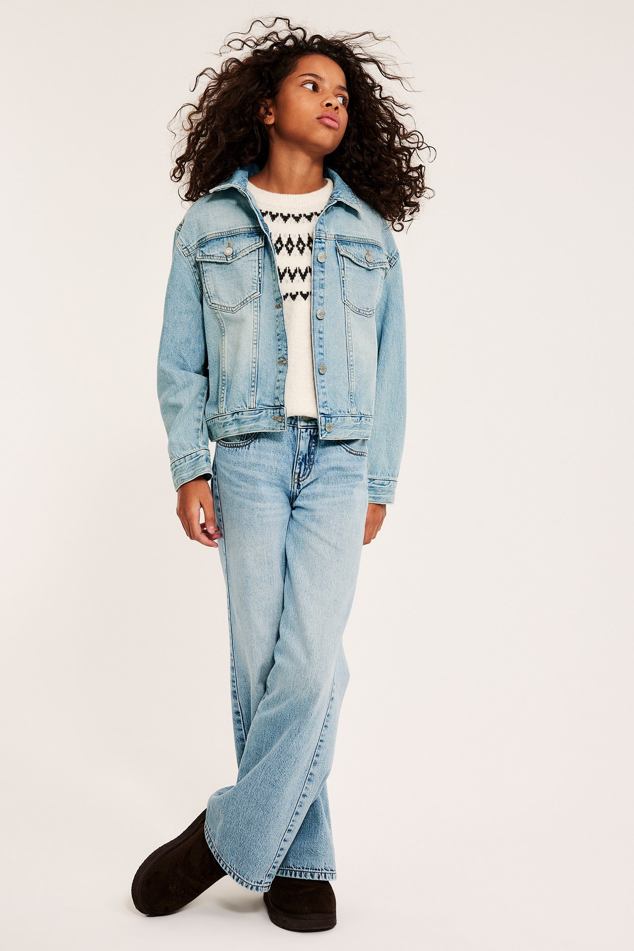 Edestä: Lapsi yllään vaaleansininen farkkutakki, valkoinen kuvioitu neule, yhteensopivat farkut ja tummat tossut.