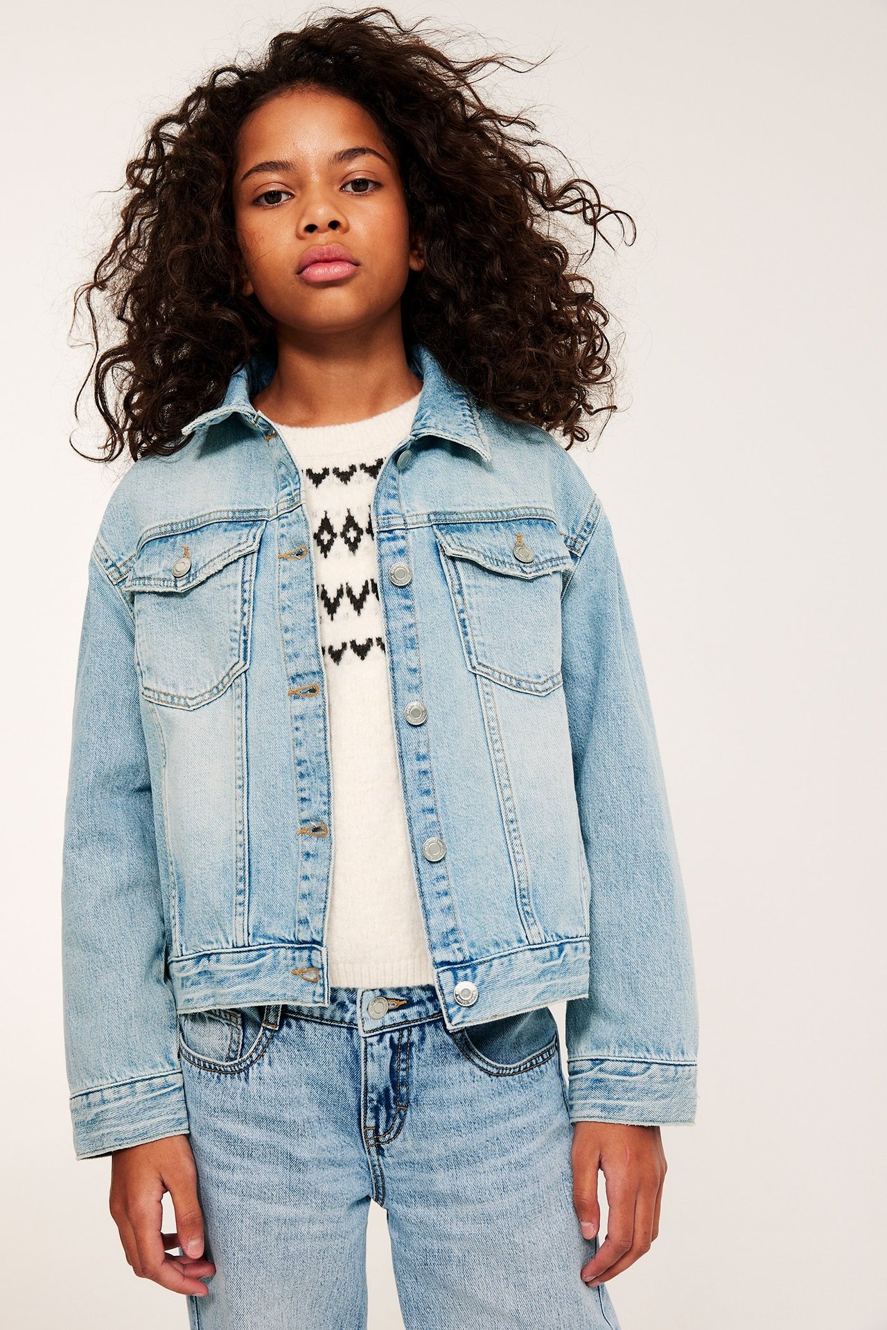 Widok z przodu: Dziecko nosi jasnoniebieską, oversizową kurtkę jeansową na białym wzorzystym swetrze i jasnoniebieskich jeansach.