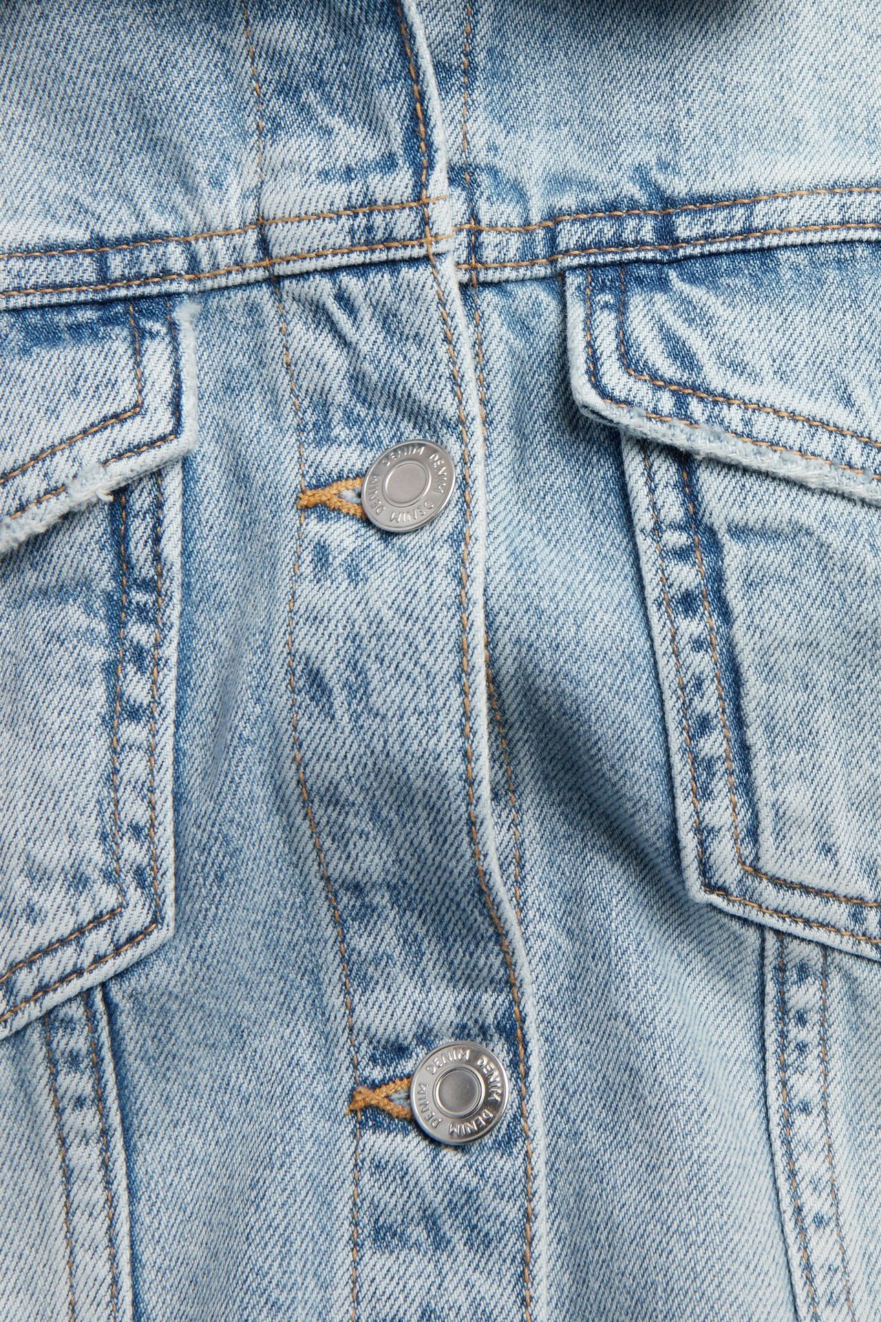 Zbliżenie na jasnoniebieską kurtkę jeansową ze srebrnymi guzikami, brązowymi szwami i kieszeniami na piersi.