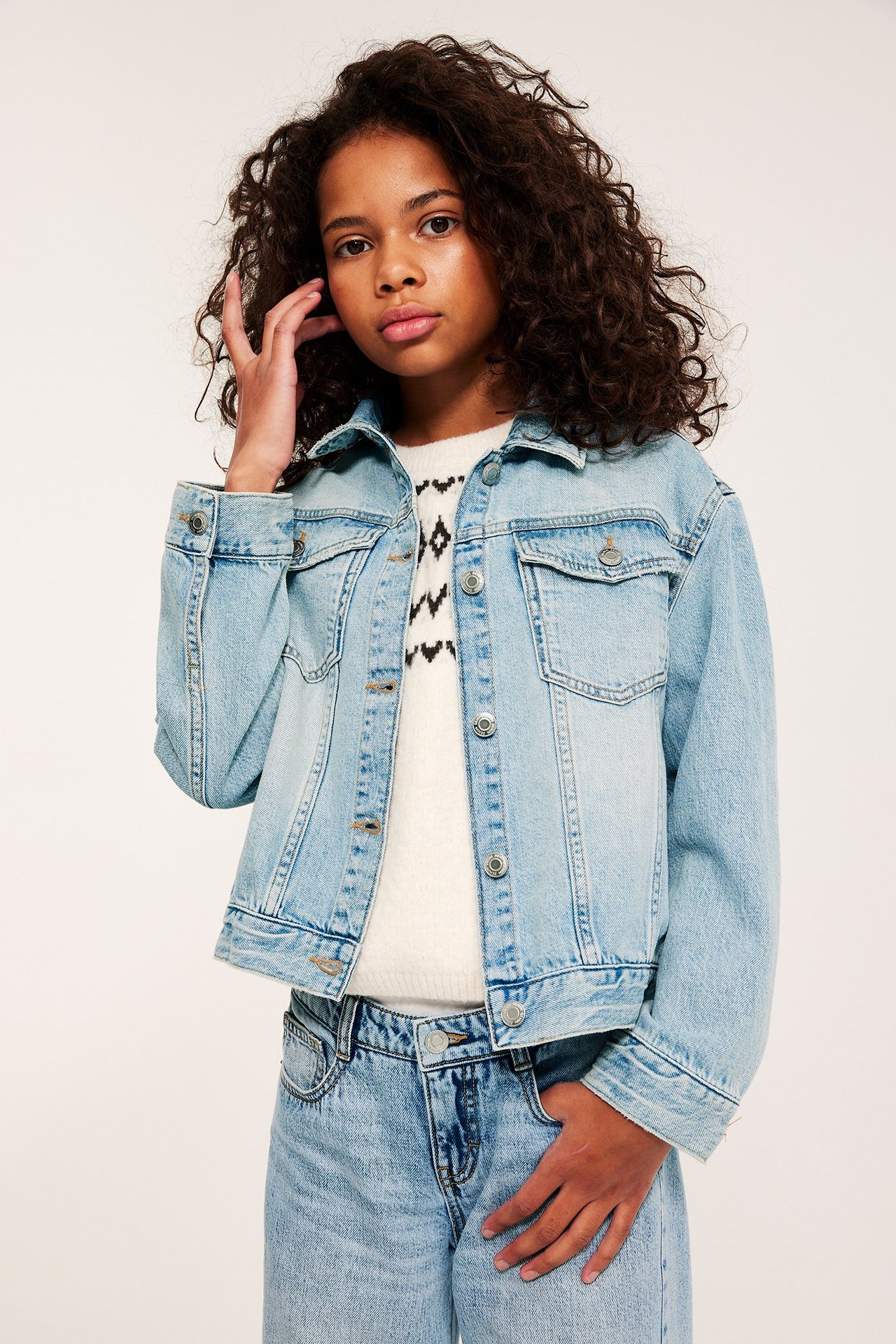 Z przodu: dziecko w jasnej, obszernej kurtce dżinsowej, na białym wzorzystym swetrze i dżinsach.