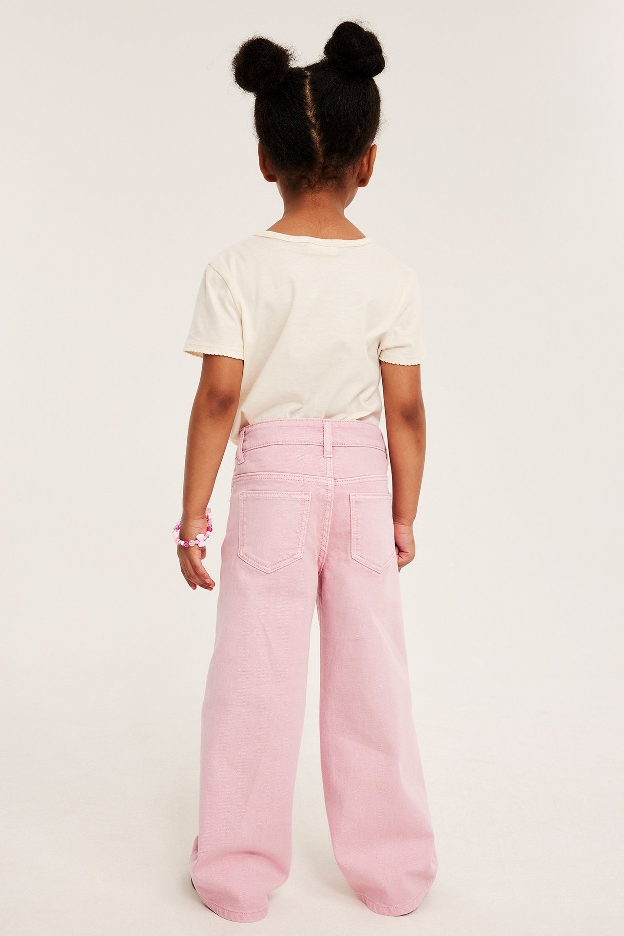 Bakfra: Barn i lys rosa vide bomullstwilljeans, beige t-skjorte, fargerikt perlearmbånd.