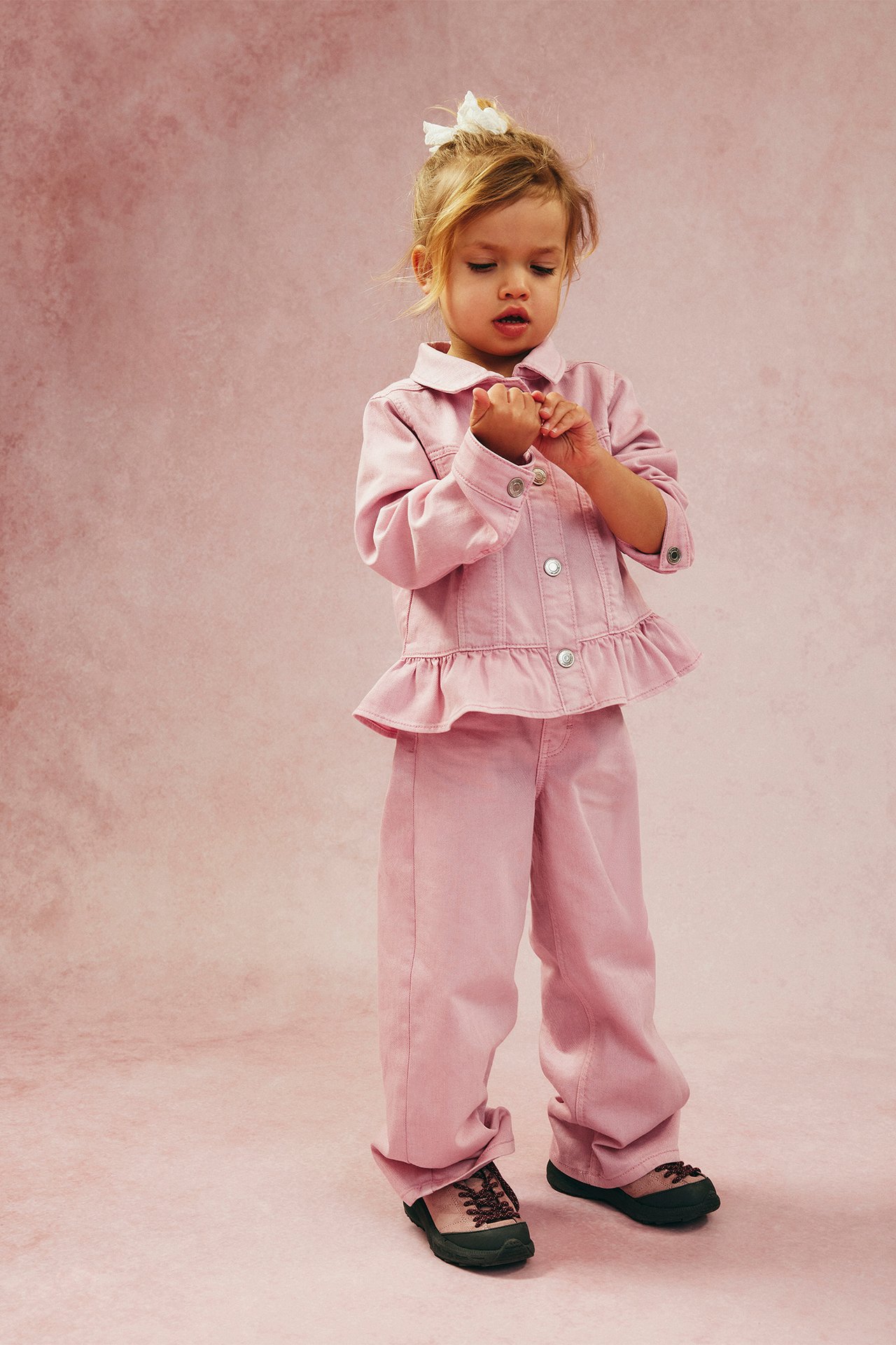 Edestäpäin lapsi vaaleanpunaisissa leveissä puuvillatwillifarkuissa, yhteensopivassa peplum-takissa ja tummissa kengissä.