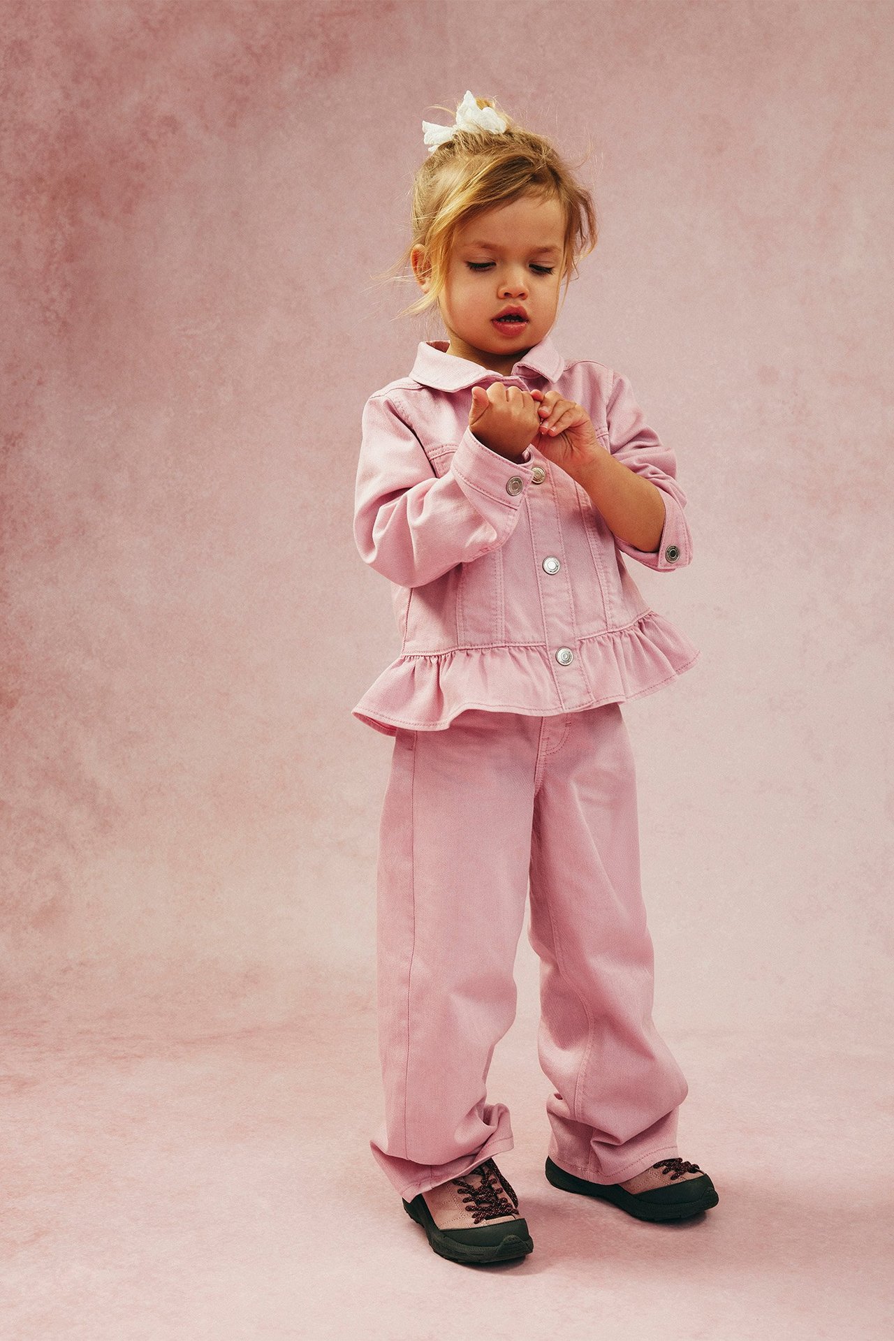 Widok z przodu dziecka w jasnoróżowych szerokich dżinsach, pasującej kurtce z baskinką i ciemnych butach.