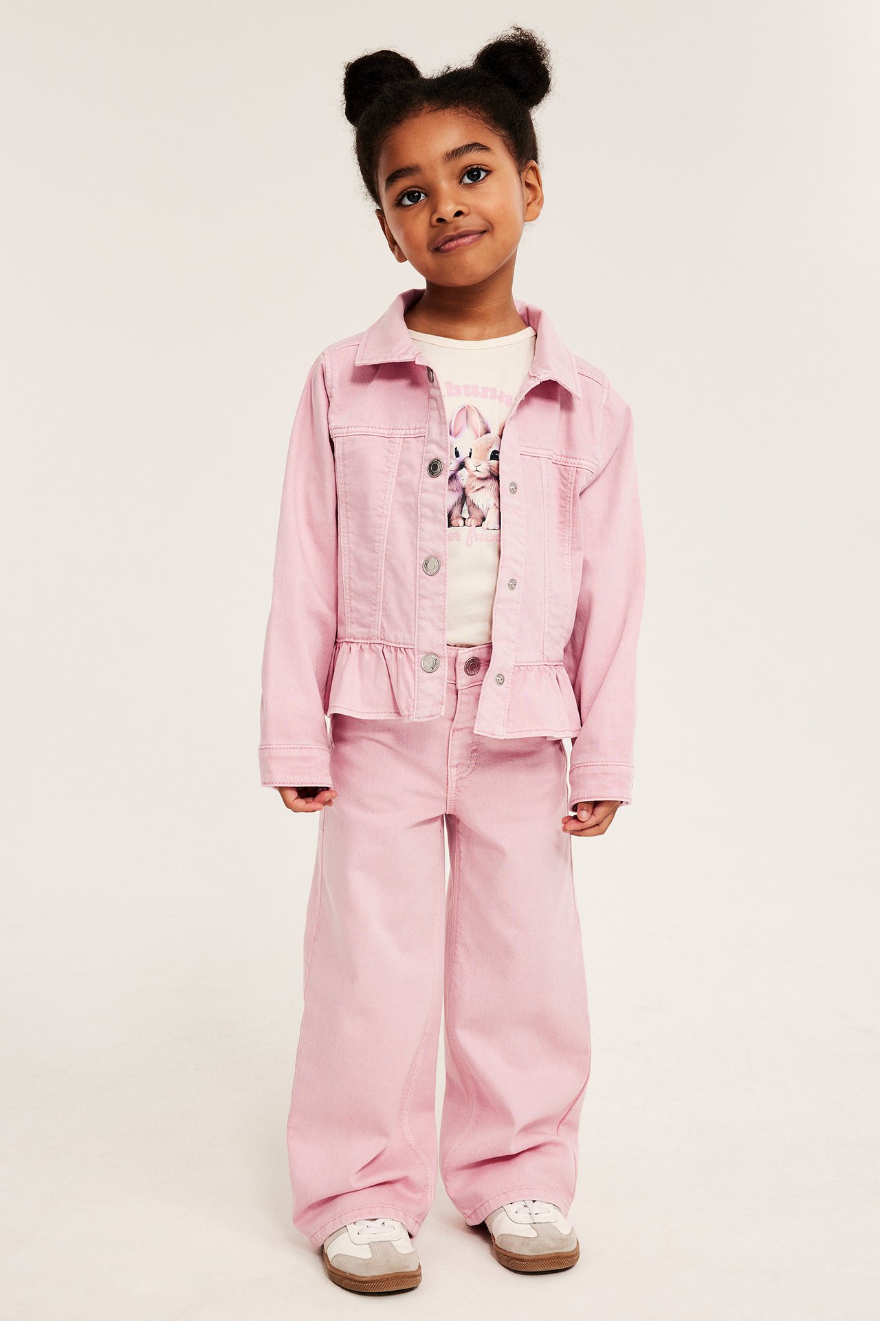 Forfra, et barn i lyserosa vide bomullstwill-jeans, matchende dongerijakke, en kanin-t-skjorte og joggesko.