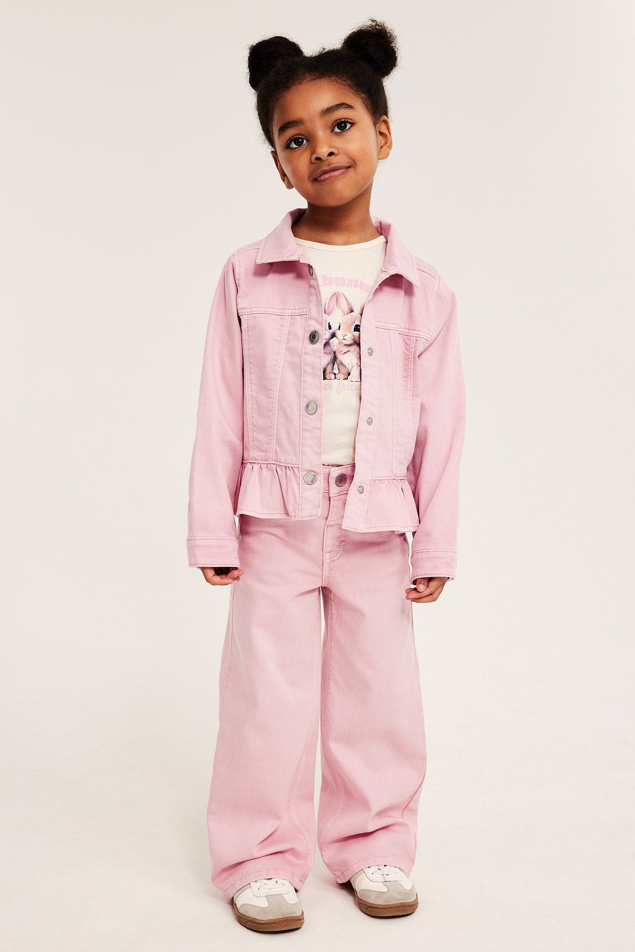 Framifrån, ett barn i ljusrosa vida bomullstwill-jeans, matchande jeansjacka, en kanintryckt t-shirt och sneakers.