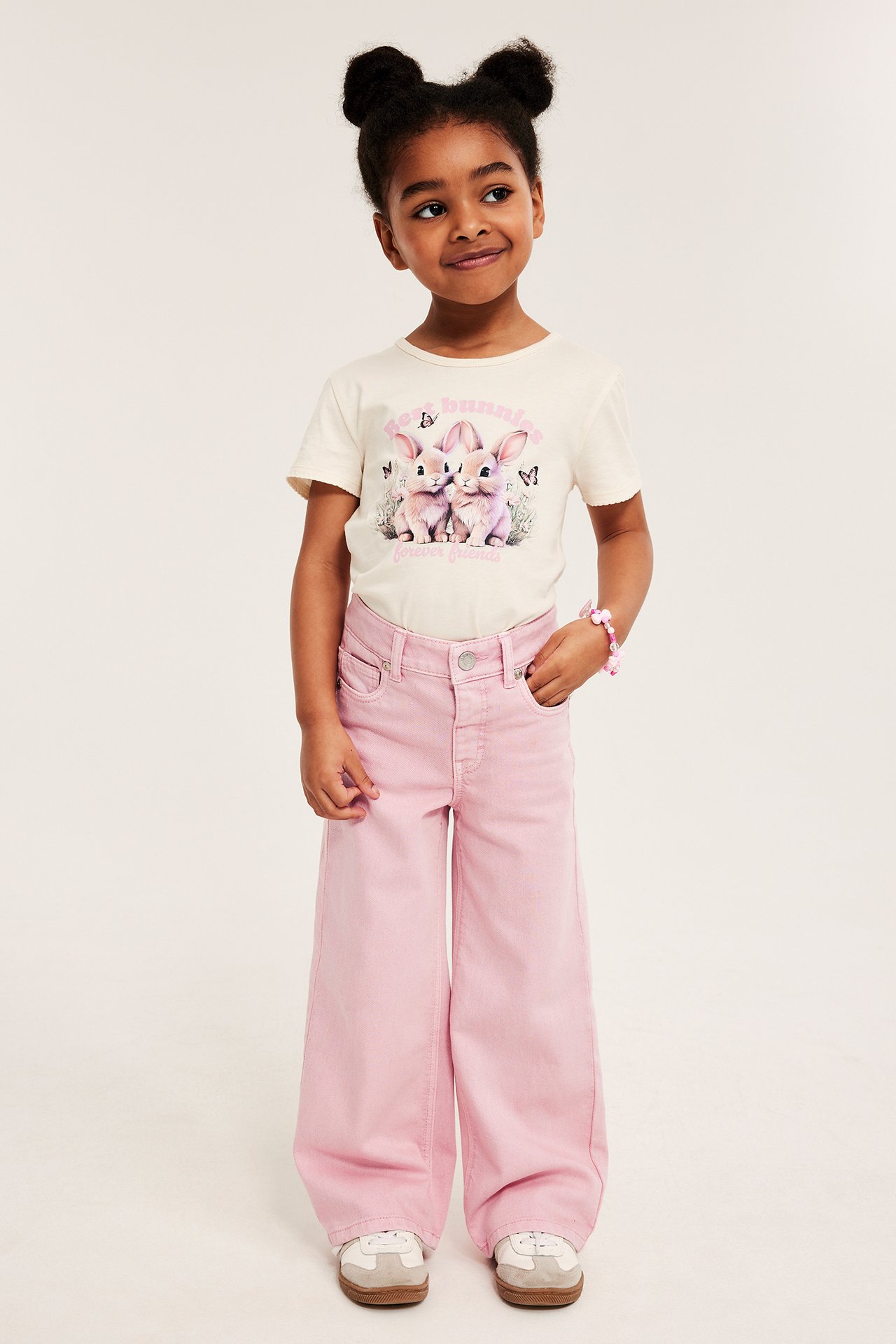 Frontalvisning: Barn iført lyserosa vide bomullsjeans med trykknapp, en kremfarget t-skjorte og hvite joggesko.