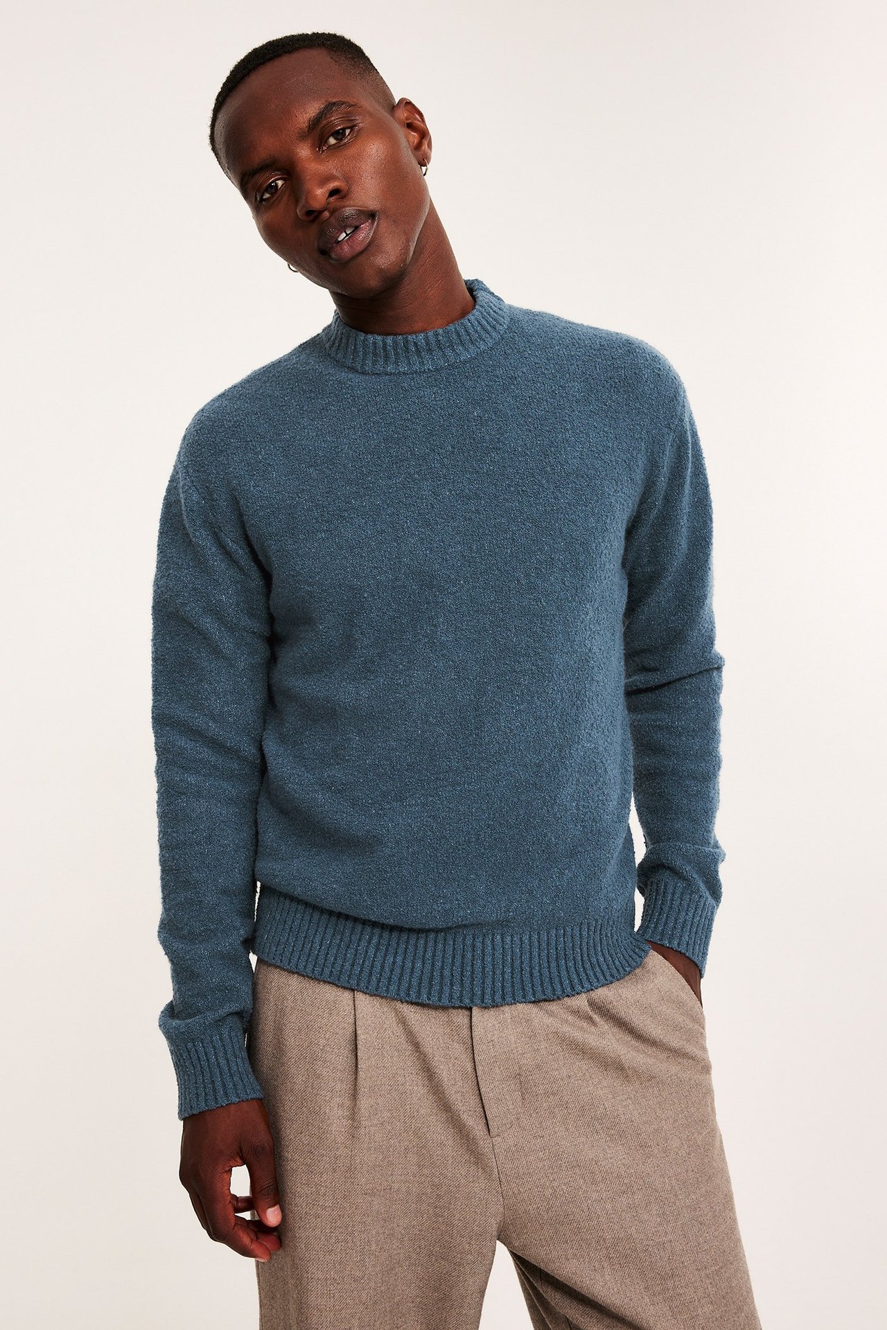 Frontbilde av en mann i en blå strikket genser med rund hals og lysebrune plisserte bukser.