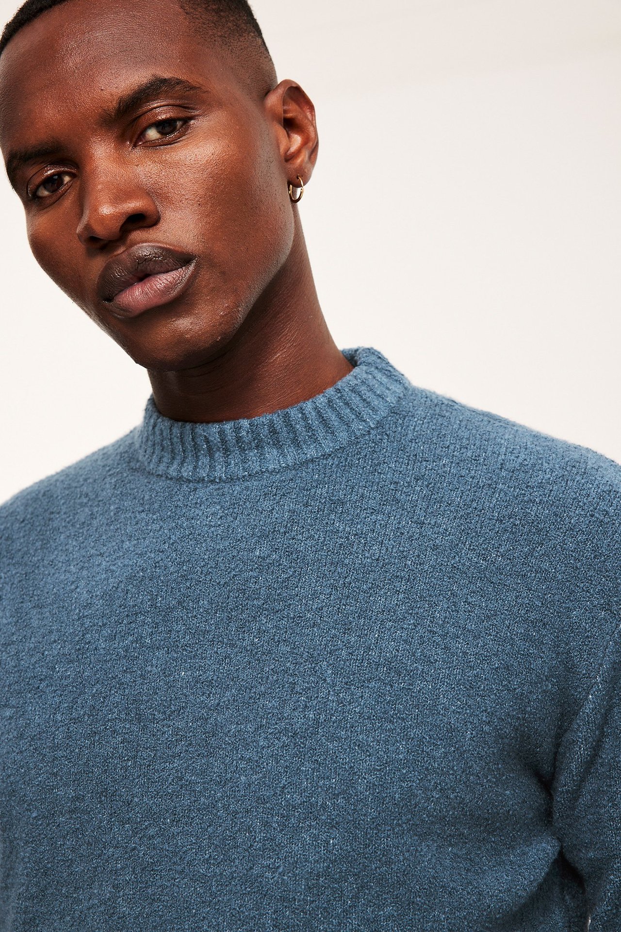 Nærbilde forfra av en mann i blå ensfarget strikket genser med halvpolo og liten gulløredobb.