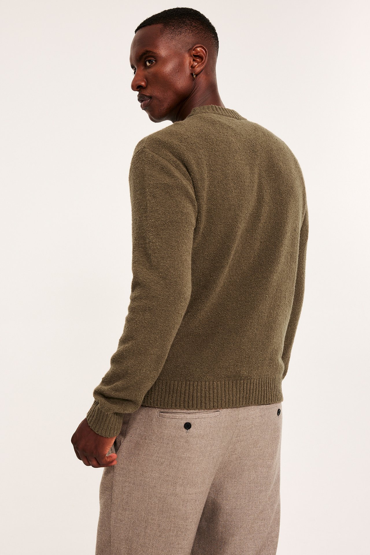 Mężczyzna tyłem w lewo, ubrany w oliwkowozielony sweter dzianinowy i jasnobrązowe spodnie.