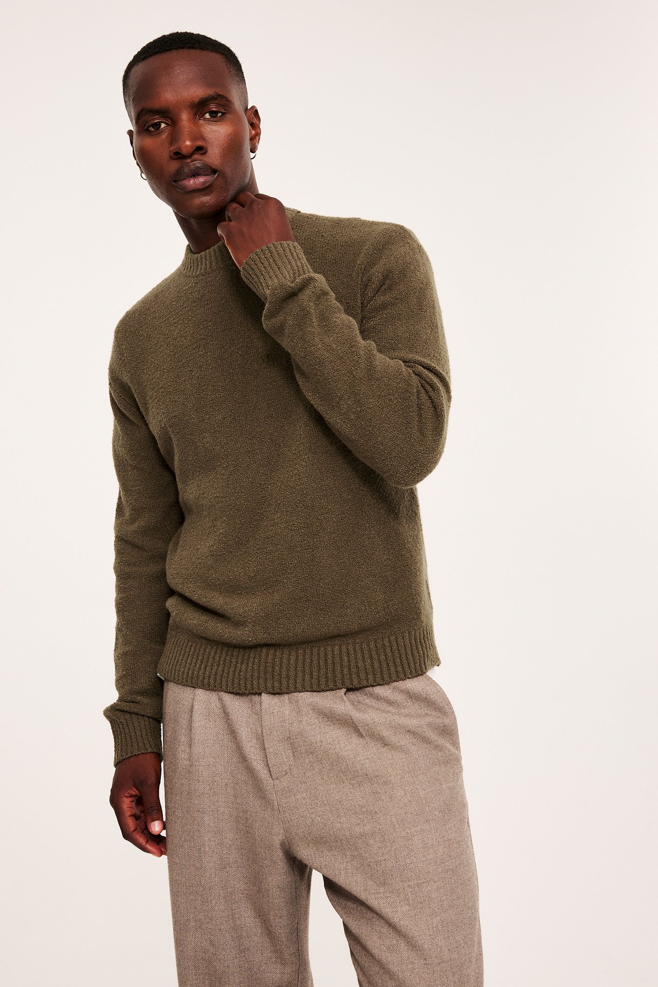 Frontvisning av en mann i en ensfarget olivengrønn strikkegenser med rund hals og lysebrune plisserte bukser.