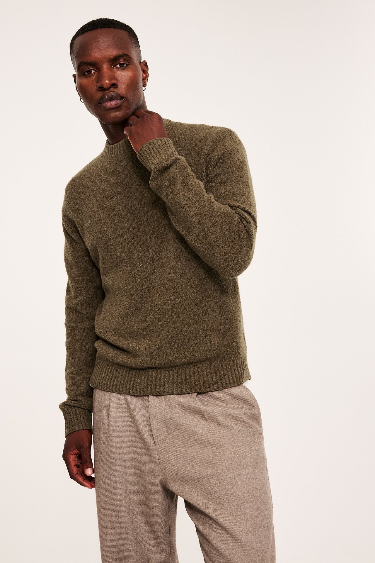 Frontvisning av en mann i en ensfarget olivengrønn strikkegenser med rund hals og lysebrune plisserte bukser.