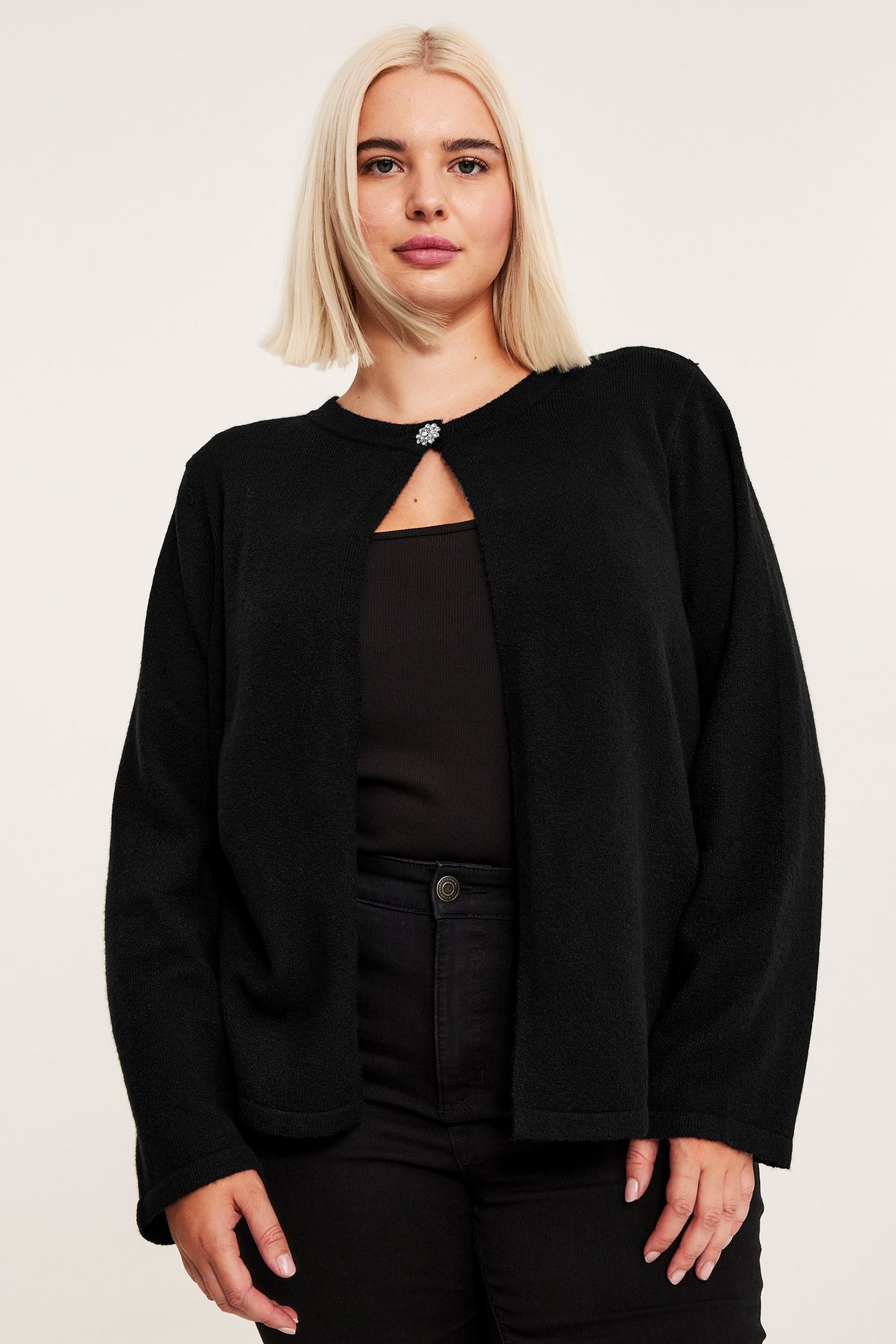 Naisen etukuva, yllään musta neuletakki strassinapilla, mustan topin ja farkkujen päällä.