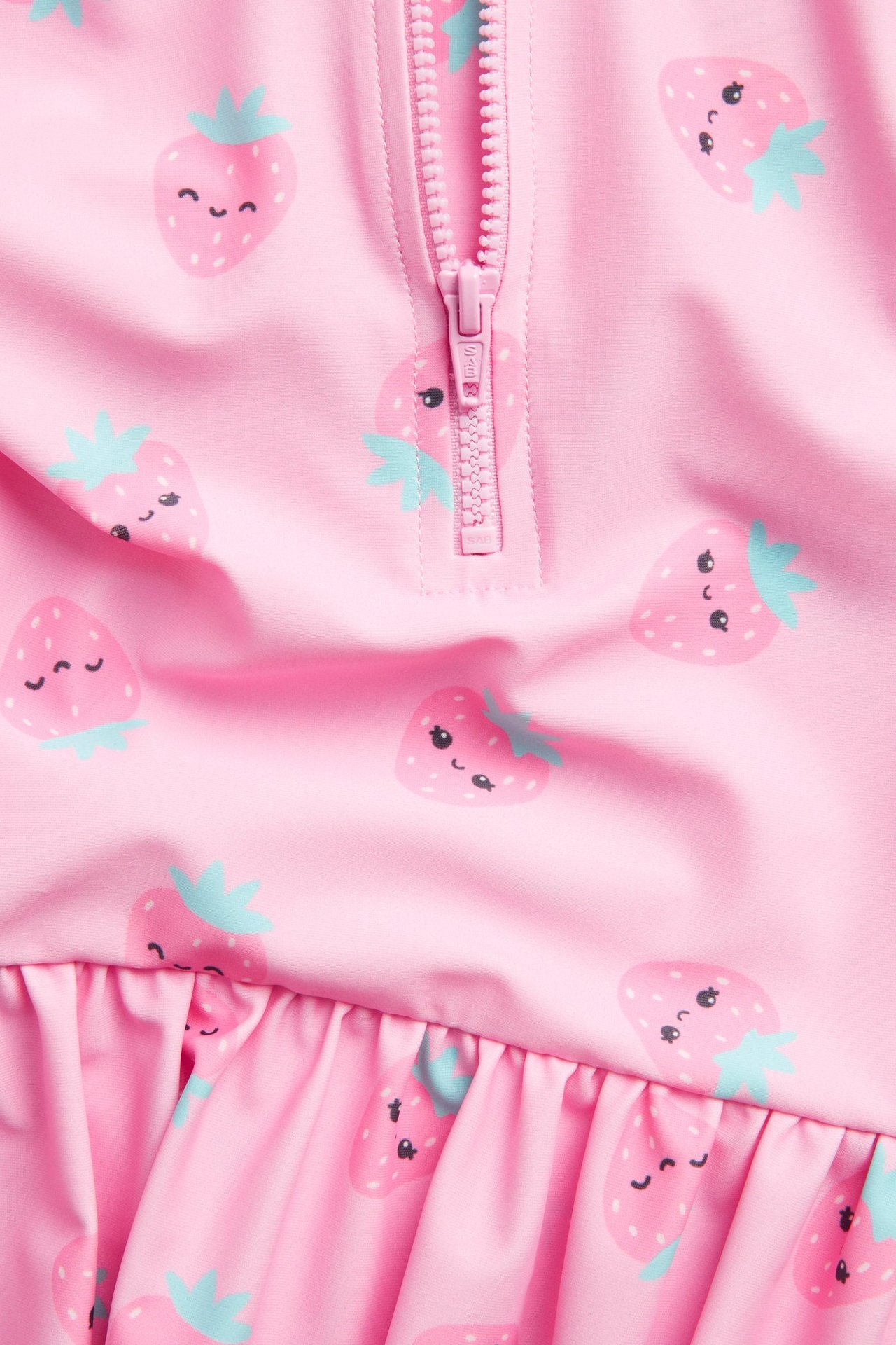 Nærbilde: lyserosa langermet UV-badedrakt med smilende jordbærmønster, glidelås og rysjekant.