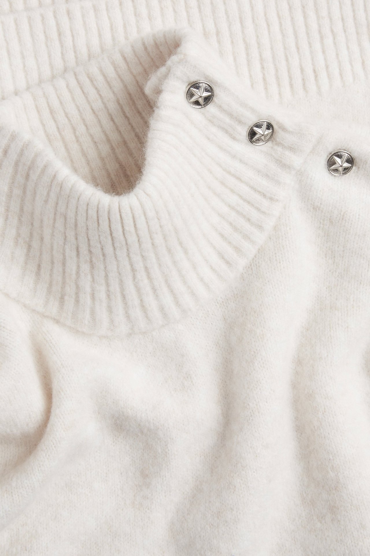 Zbliżenie białego swetra z golfem, prążkowanym kołnierzem i trzema srebrnymi guzikami-gwiazdkami.