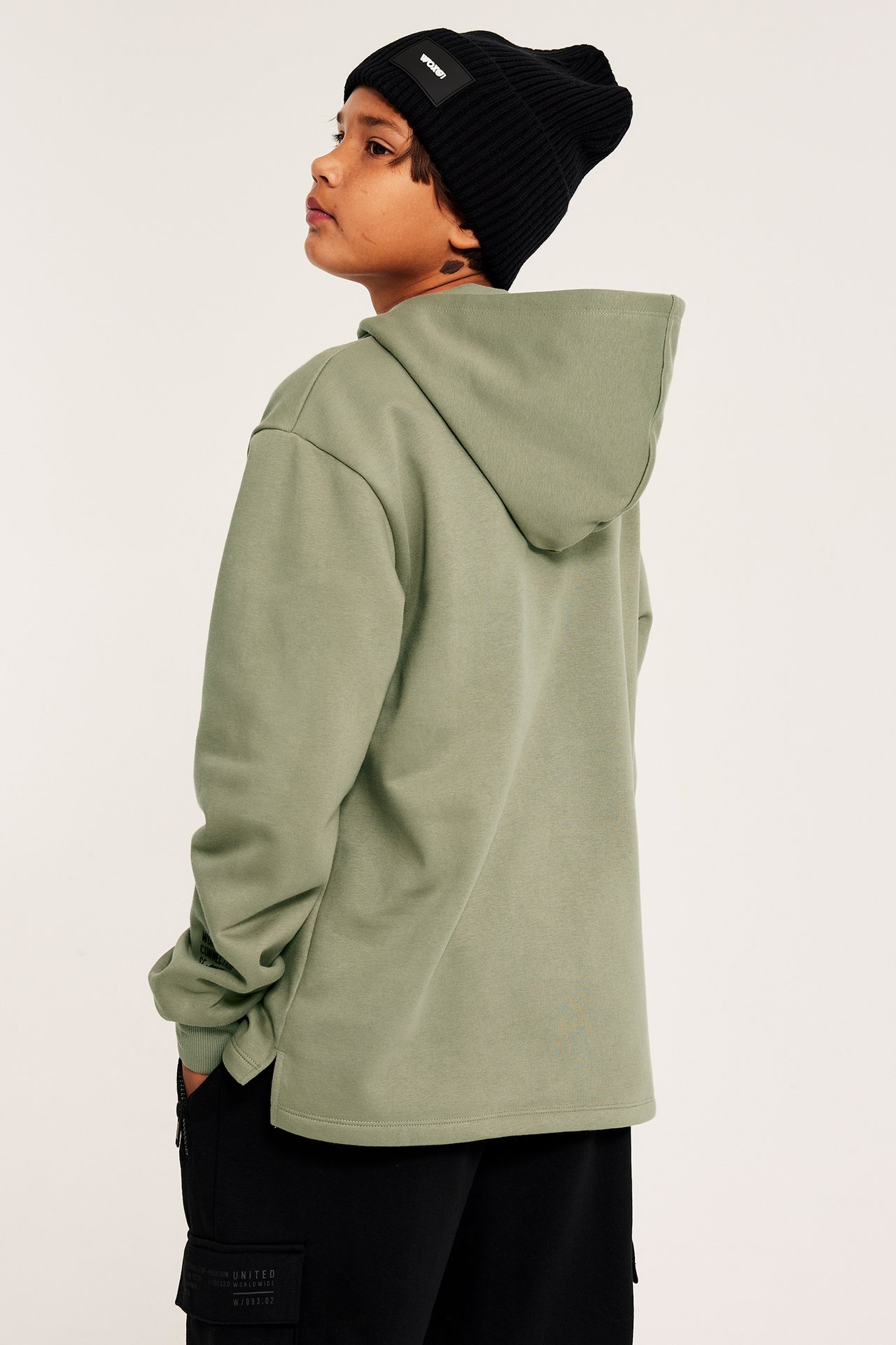 Sidovy av ett barn i en grön bomullsblandad hoodie, svarta byxor och svart ribbad mössa.