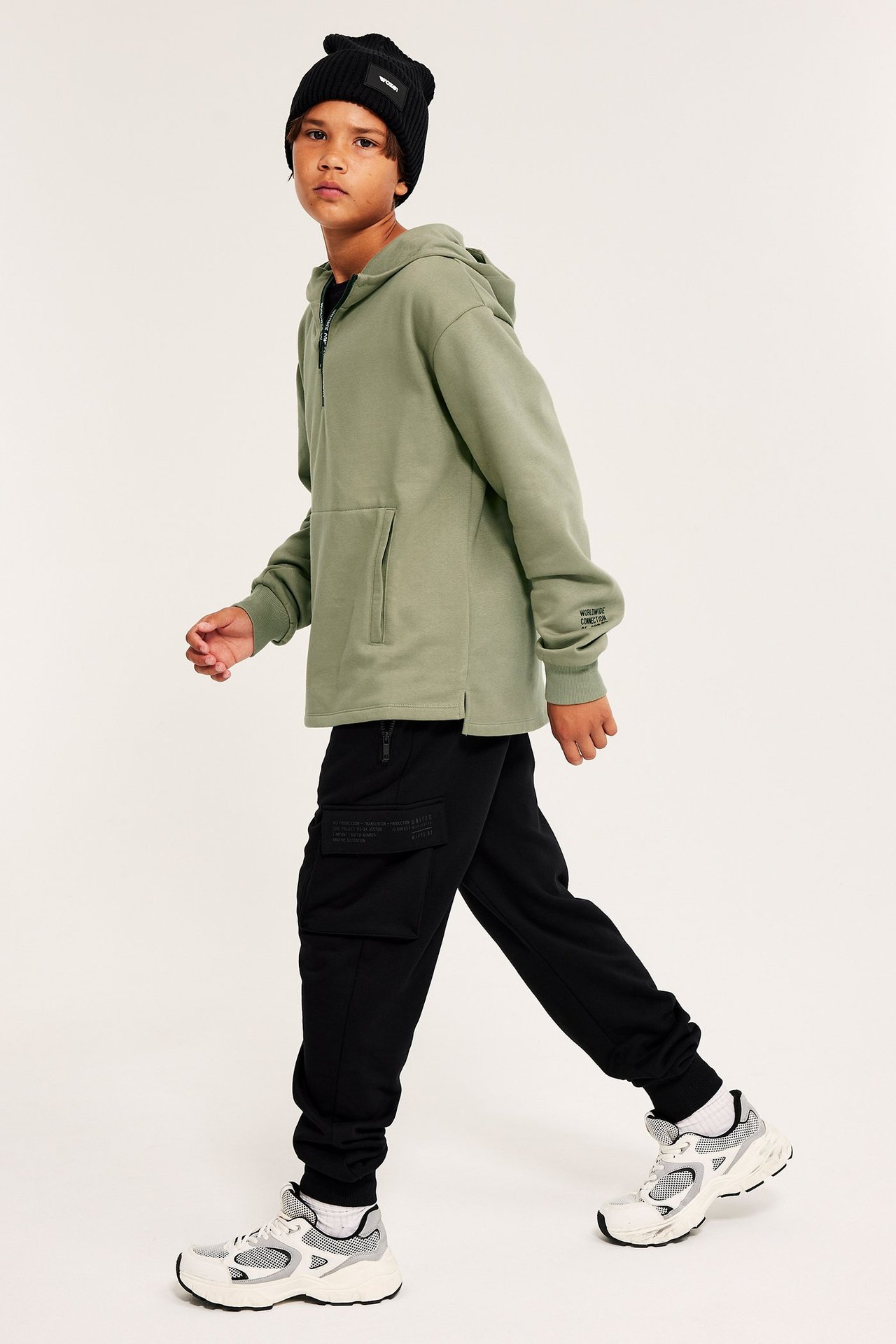 Widok z boku dziecka w zielonej bluzie z zamkiem, czarnej czapce, czarnych spodniach cargo i białych sneakersach.