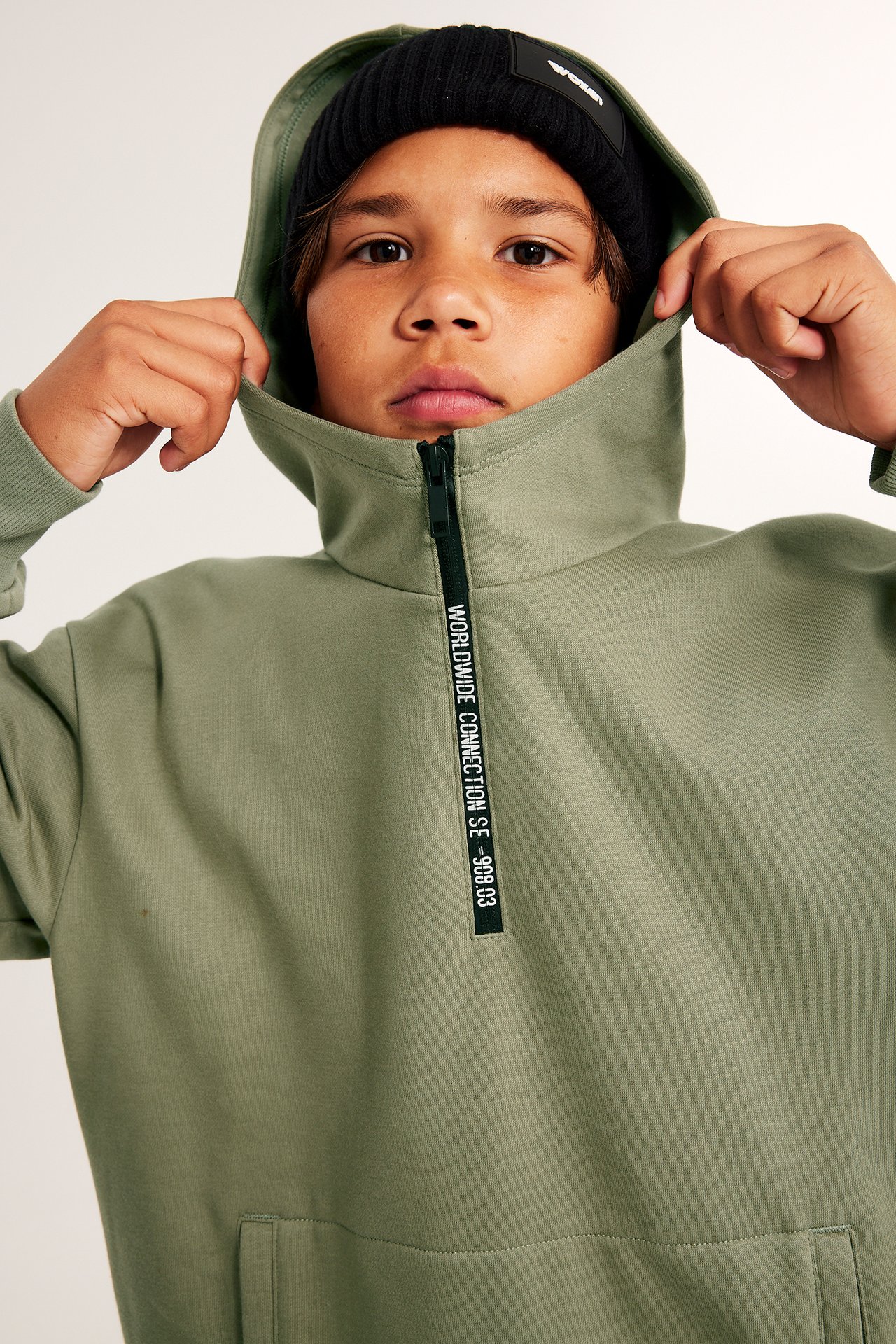 Närbild framifrån på ett barn i grön huvtröja med textdragkedja och svart mössa.