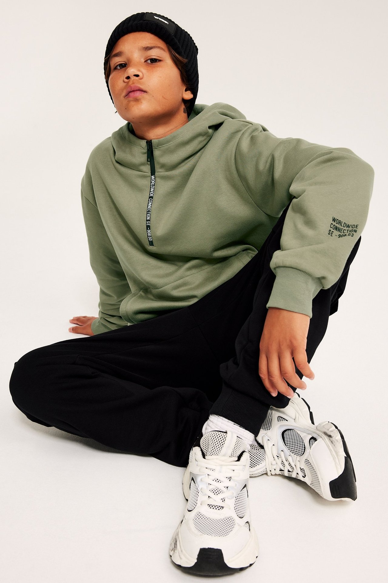 Barn framifrån: grön zip-hoodie, svarta mjukisbyxor, svart mössa och vita sneakers.