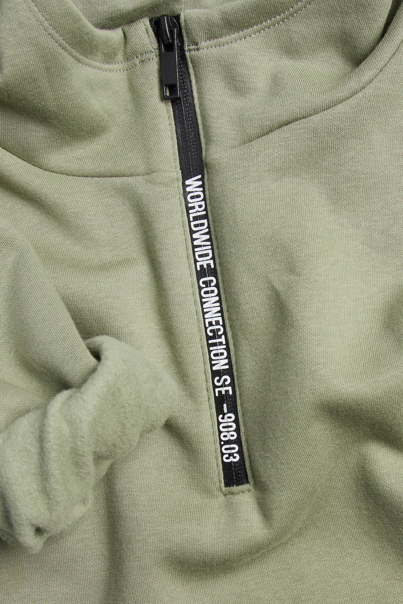 Närbild av en grön barnhoodie, svart dragkedja med texten 'WORLDWIDE CONNECTION SE -908.03'.
