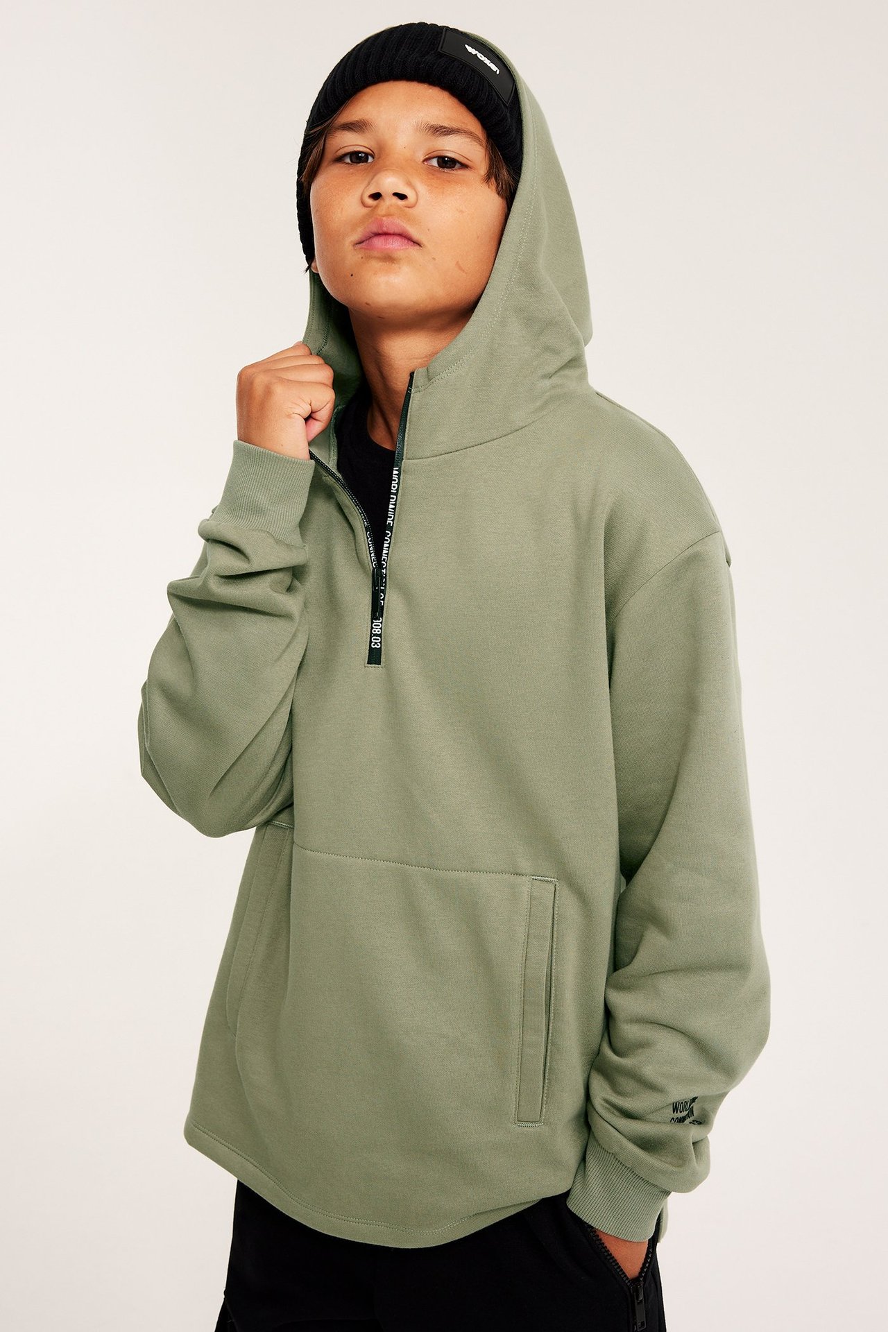 Framifrån, ett barn i en salviagrön halvzip-hoodie, med svart mössa och svarta byxor.