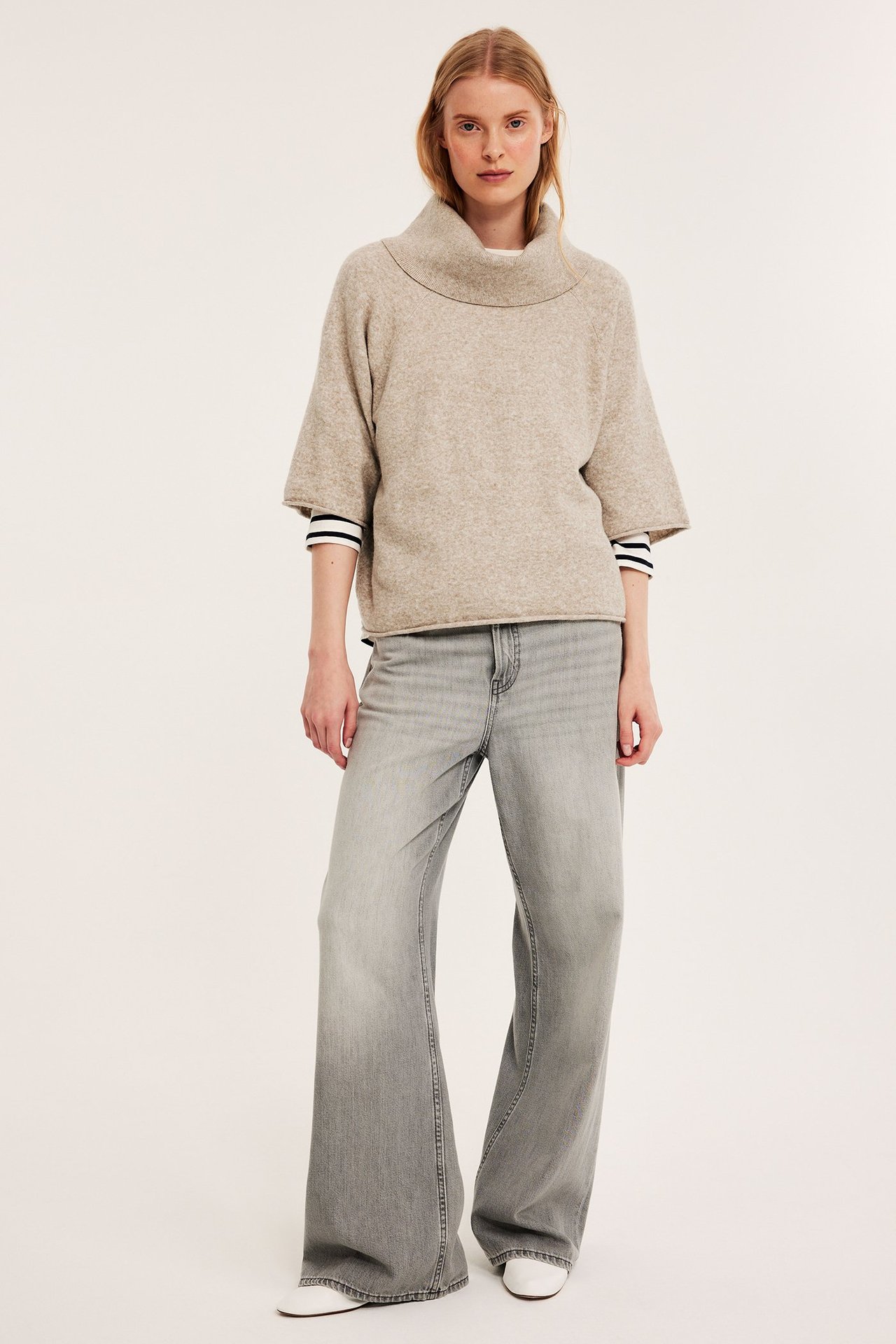 Nainen yllään beige meleerattu neulepoolo, raidallinen paita ja harmaat levenevät farkut.