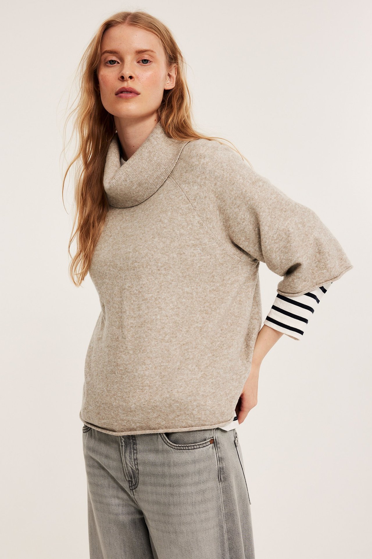 Widok z przodu: Kobieta w beżowym melanżowym swetrze z golfem, koszulce w paski i szarych dżinsach.